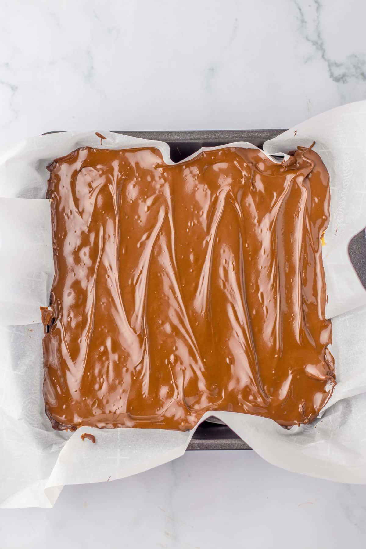 snickers fudge in a square parchment lined baking sheet ready for the fridge to chill.