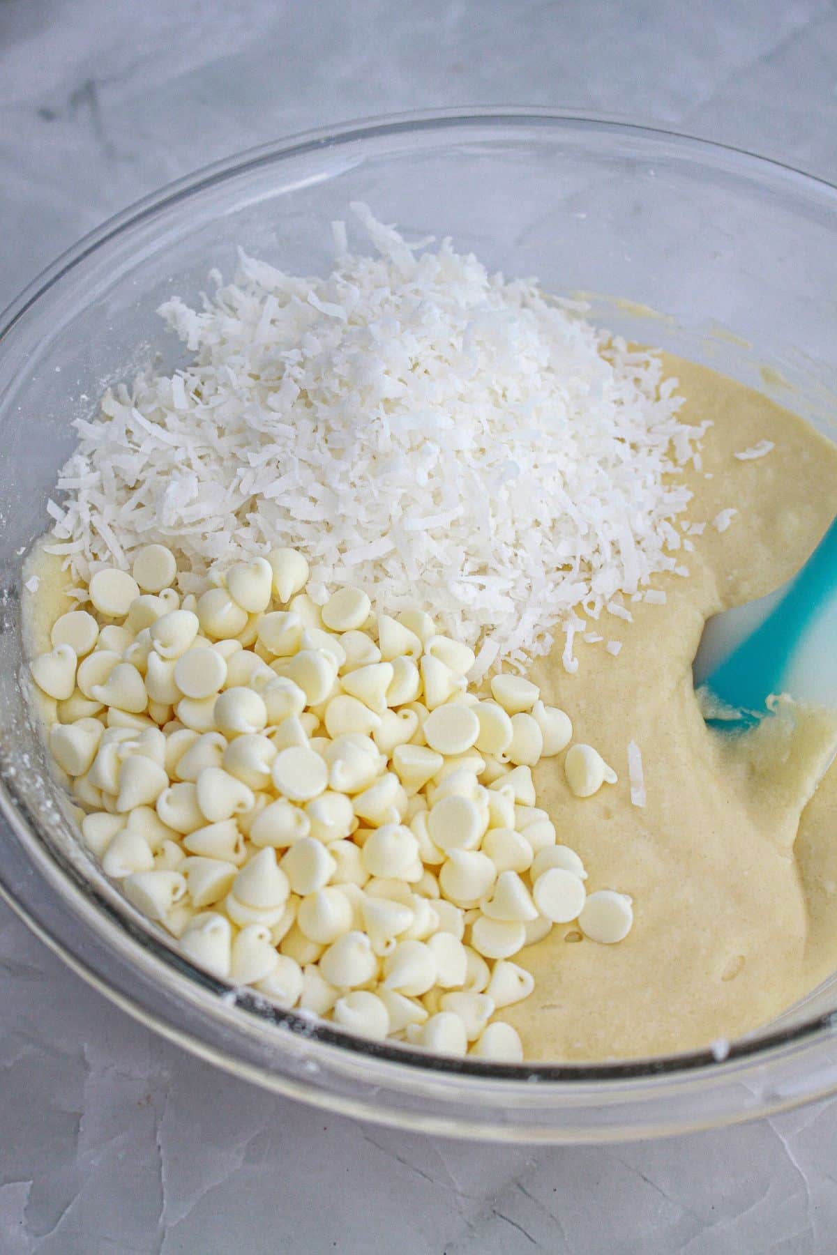 chocolate chips and coconut flakes being added to cake batter in a glass bowl.