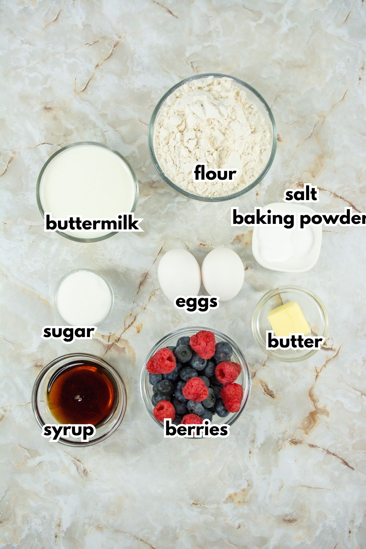 bowls of flour, buttermilk, sugar, eggs, salt, baking powder, syrup, berries and butter on a counter.