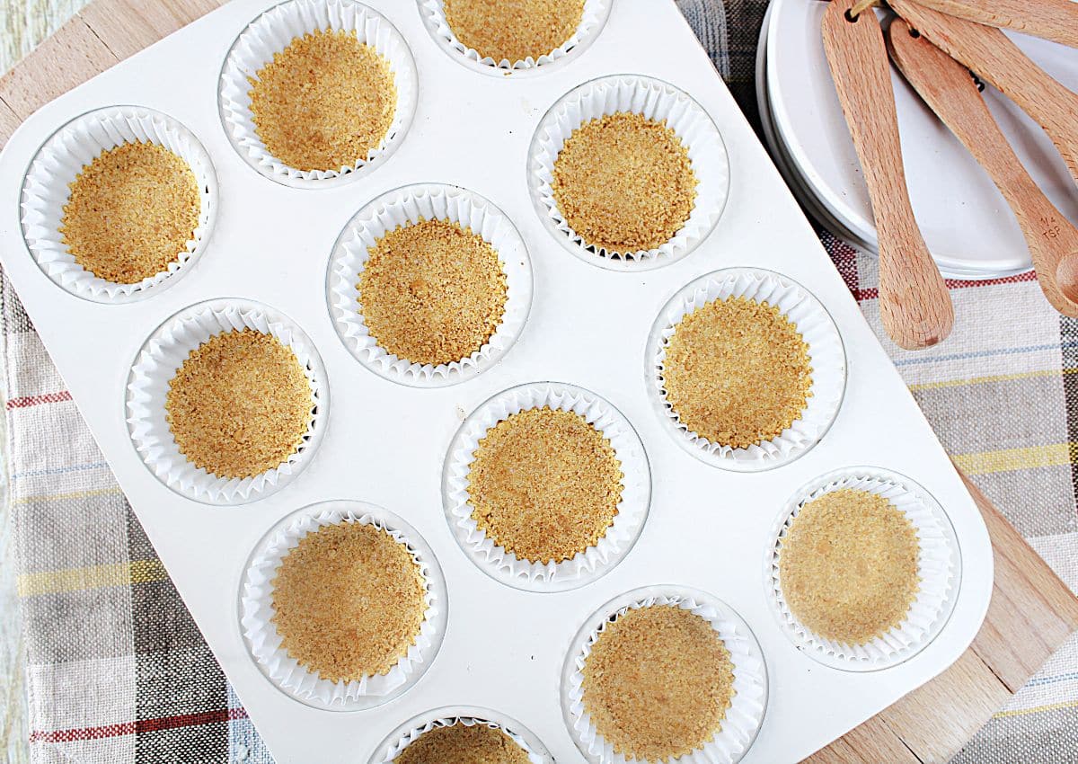 graham cracker crust being pressed into the bottom of paper lined muffin pan.