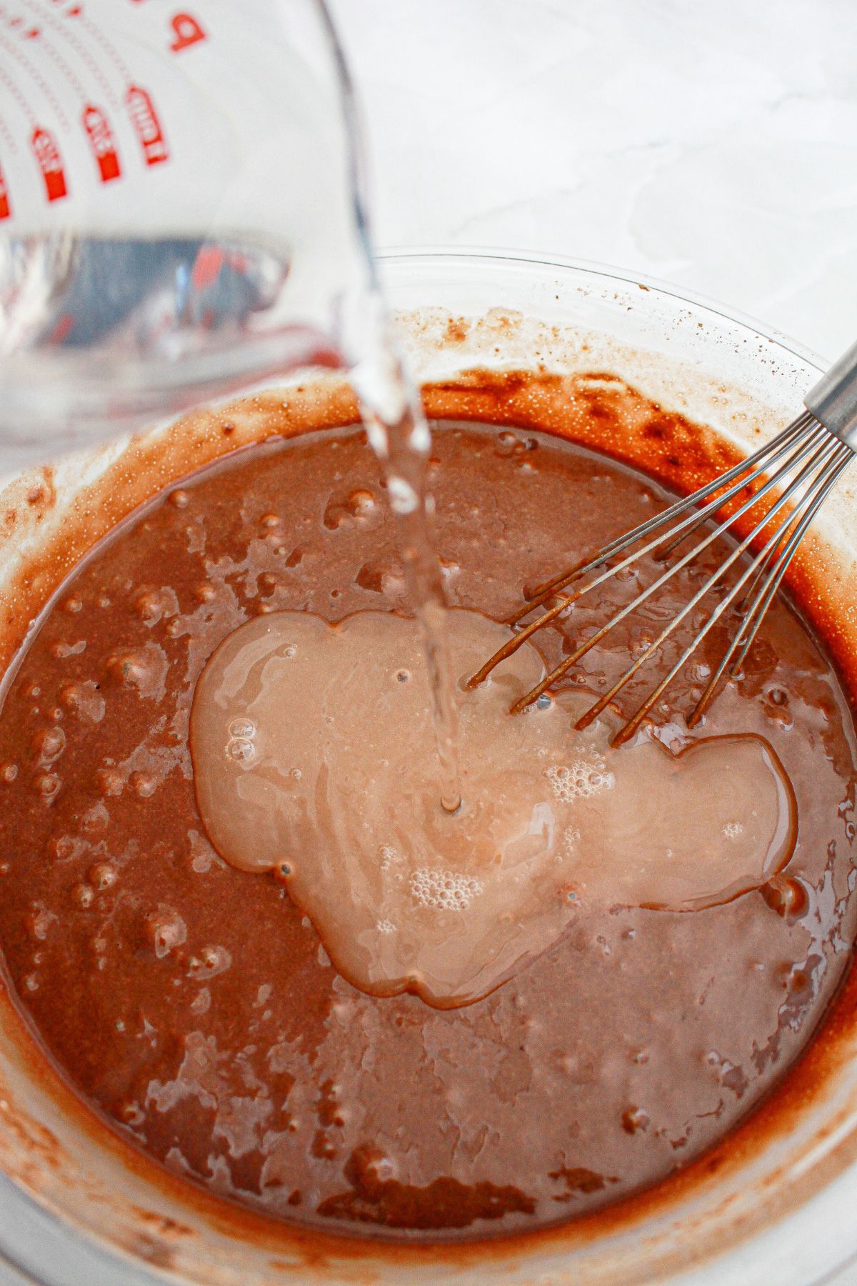 chocolate cake batter being mixed in a glass mixing bowl with a whisk.