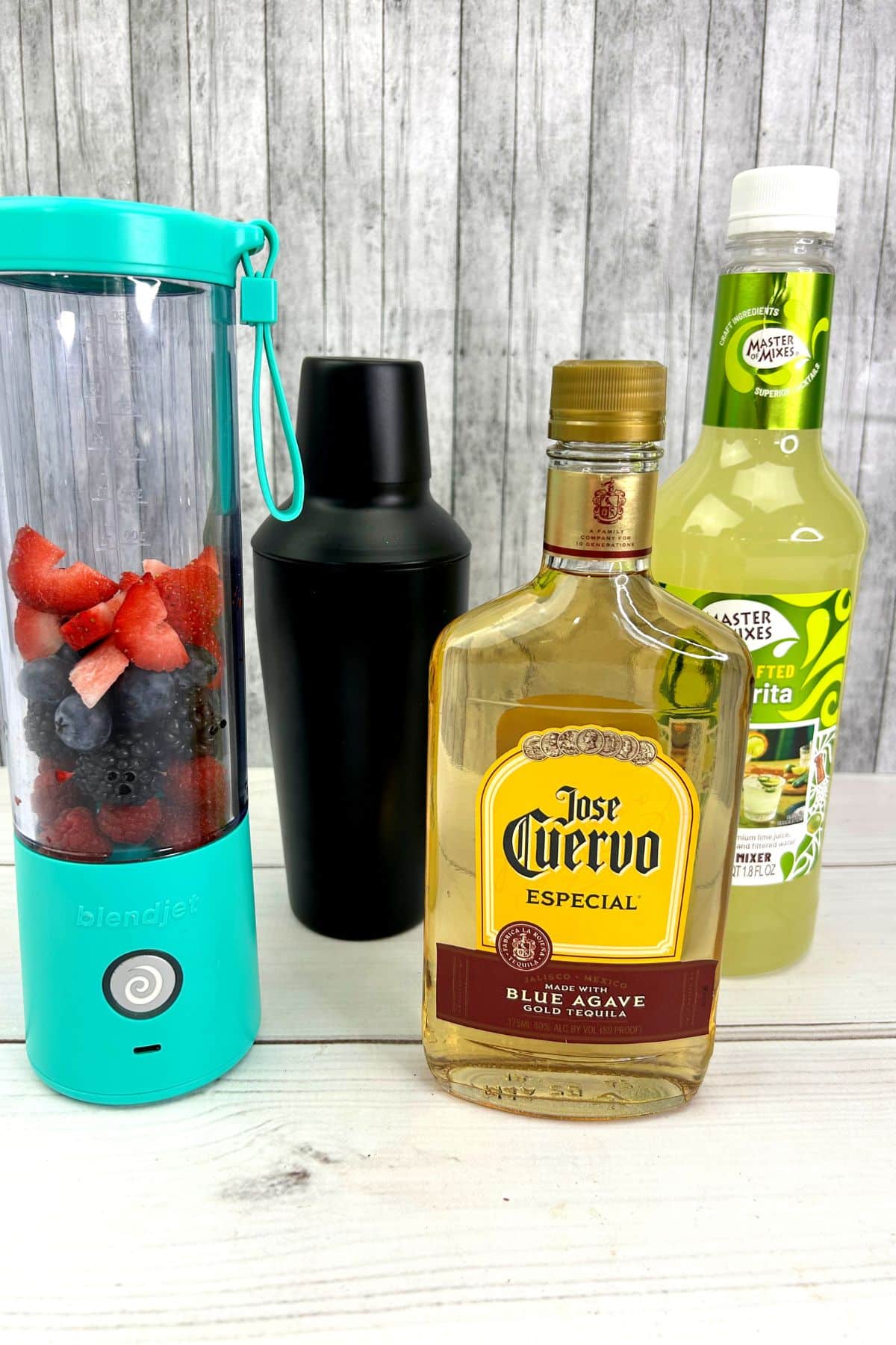 bottles of tequila, sweet and sour mix, a cocktail shaker and a blend jet full of berries on a counter.