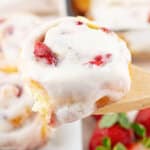 glazed strawberry cinnamon roll held in the air on a wooden spoon.