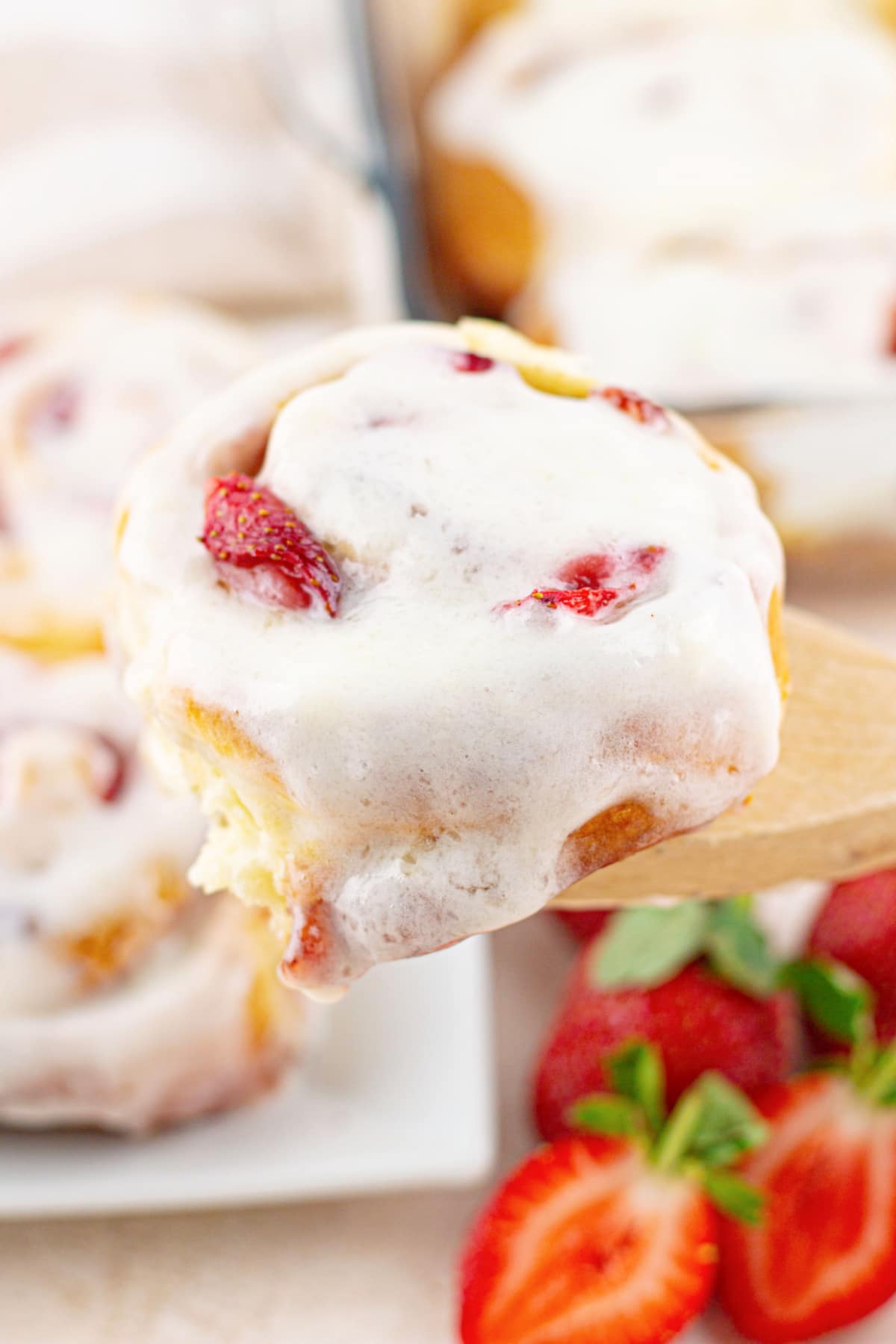 glazed strawberry cinnamon roll held in the air on a wooden spoon.