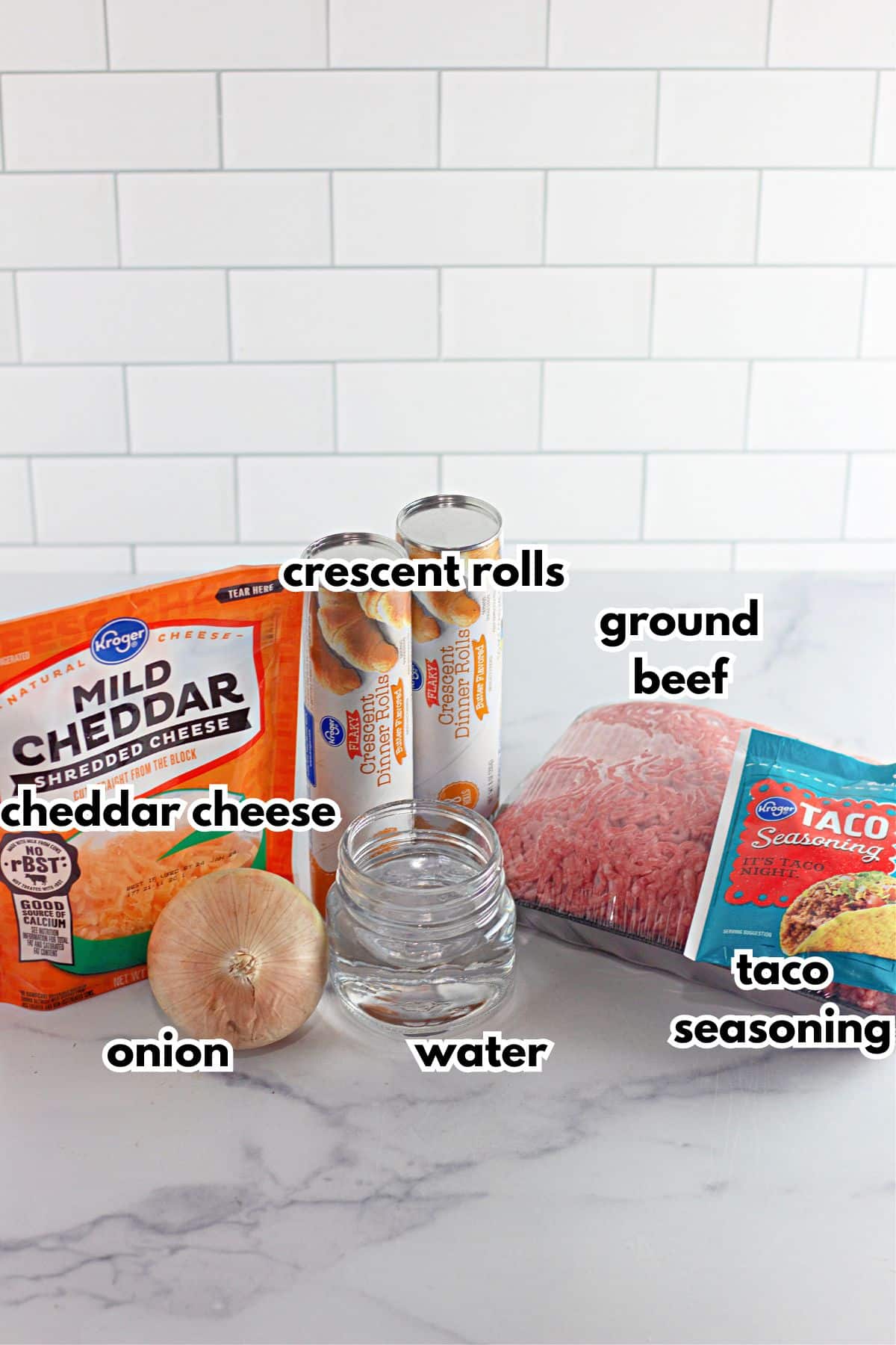 bag of cheddar cheese, two cans of crescent rolls, onion, water, taco seasoning packet, and a package of ground beef on a counter.