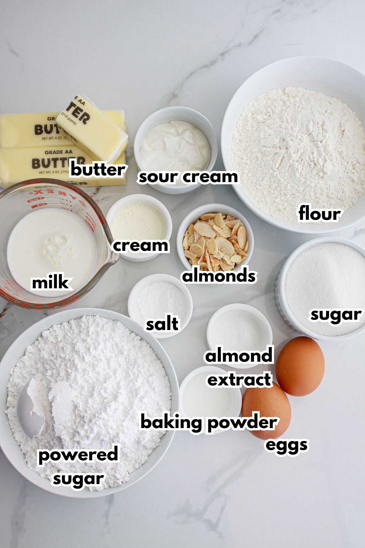 bowls of butter, sour cream, flour, milk, cream, almonds, salt, almond extract, sugar, eggs, baking powder and powdered sugar.