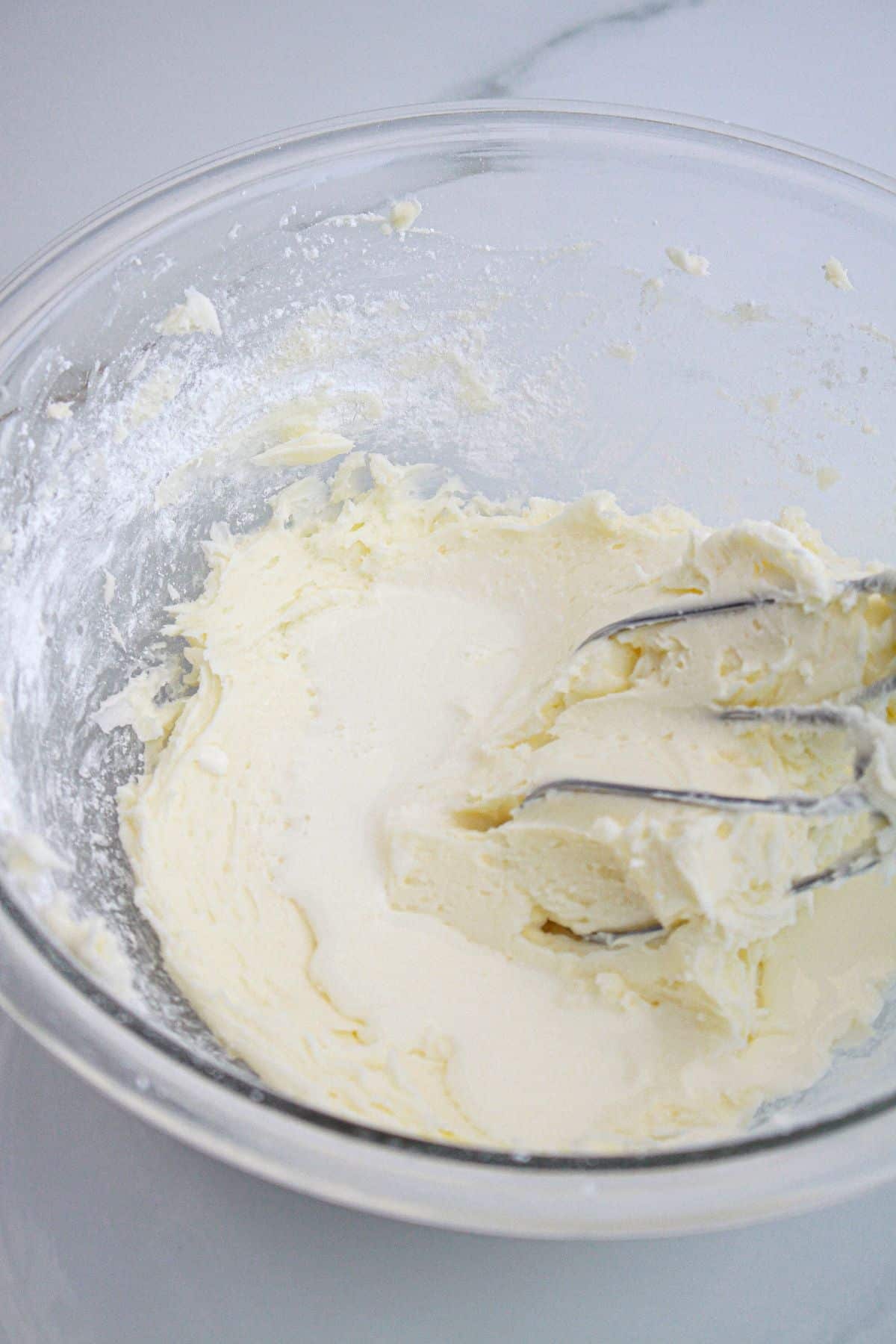 heavy cream being added to frosting in a glass mixing bowl.
