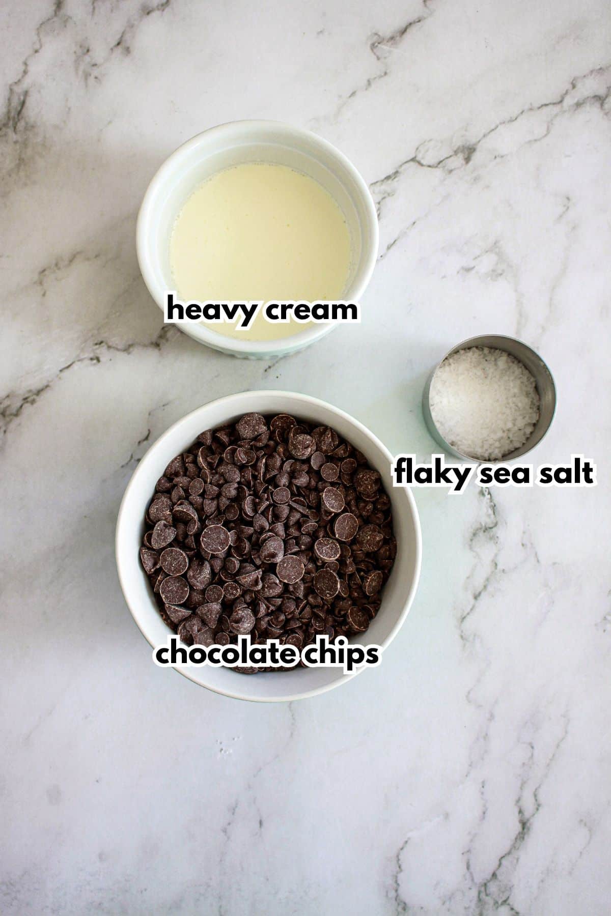 bowls of semi-sweet chocolate chips, flaky sea salt, and heavy cream on a counter.