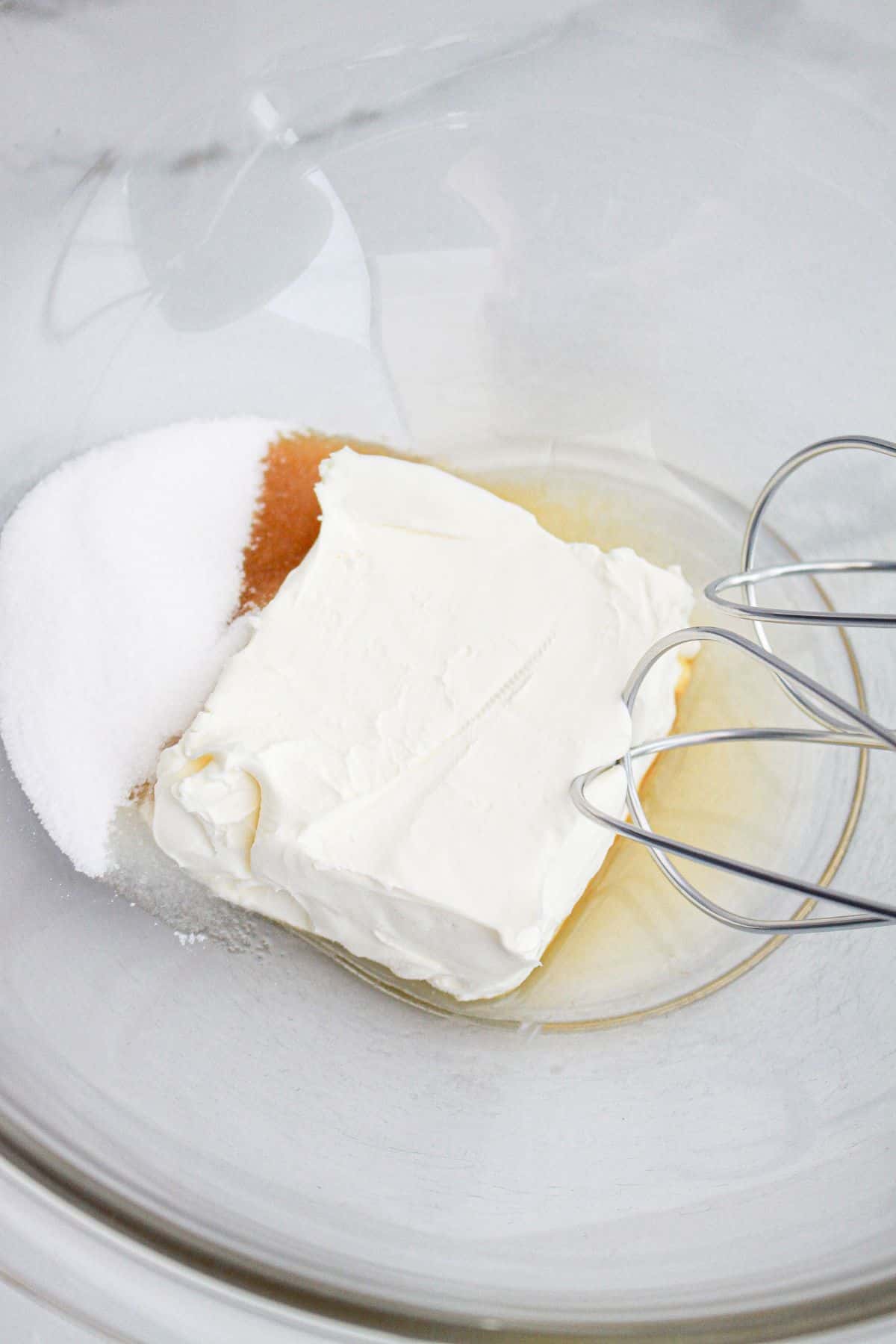 cream cheese, sugar, and vanilla being mixed in a glass mixing bowl.