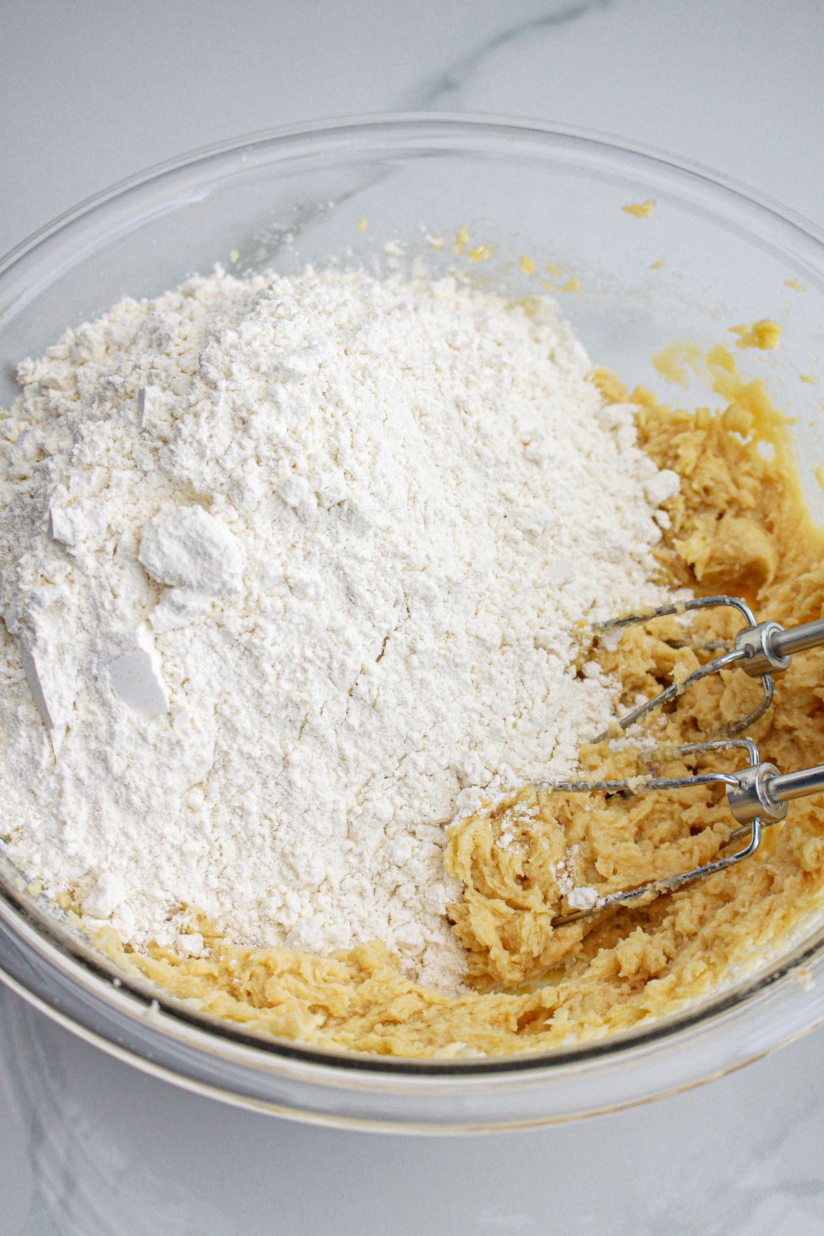 flour being added to cookie dough in a glass mixing bowl.