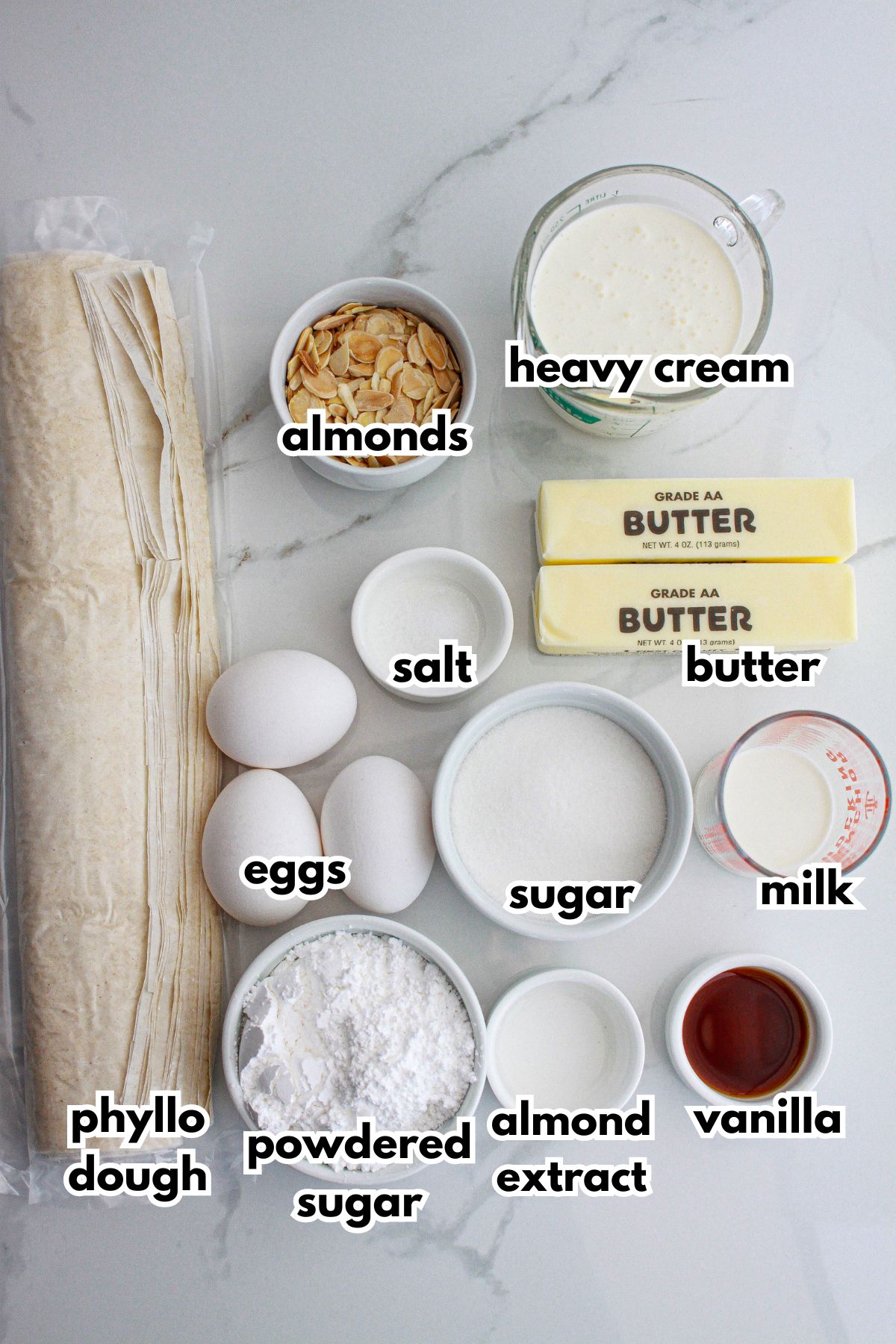 roll of phyllo dough, bowls of almonds, heavy cream, salt, butter, eggs, sugar, milk, powdered sugar, almond extract, and vanilla on a counter.