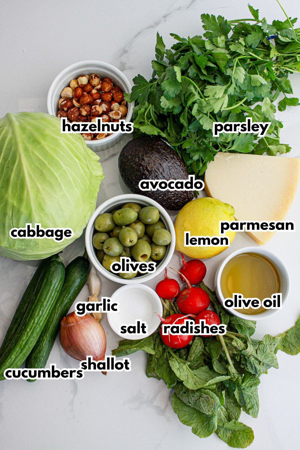 hazelnuts, parsley, cabbage, avocado, lemon, parmesan cheese, olives, garlic, salt, hallows, cucumbers, radishes and olive oil on a counter.