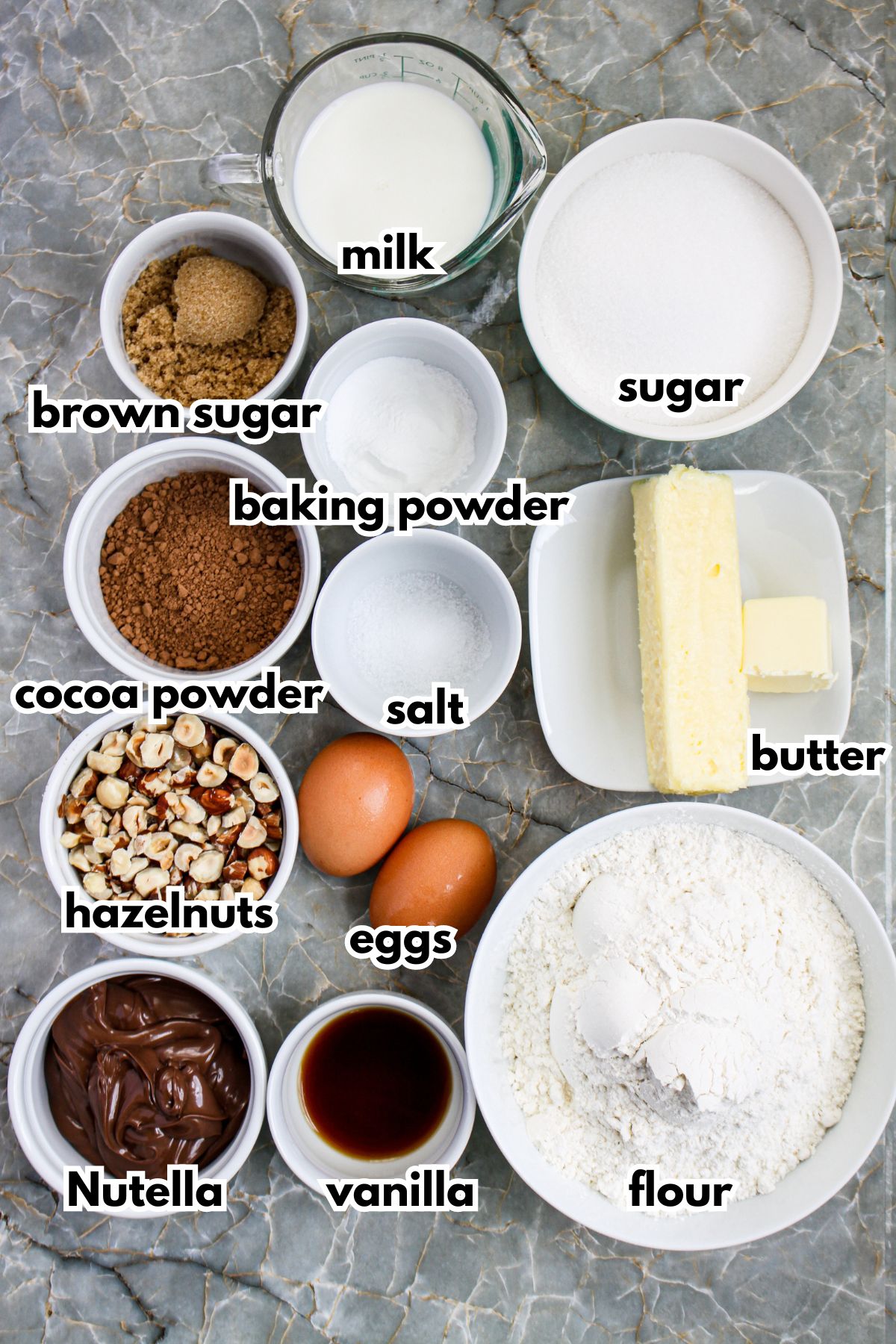 bowls of milk, brown sugar, sugar, baking powder, salt, cocoa powder, butter, hazelnuts, Nutella,, vanilla, ad flour on a counter.