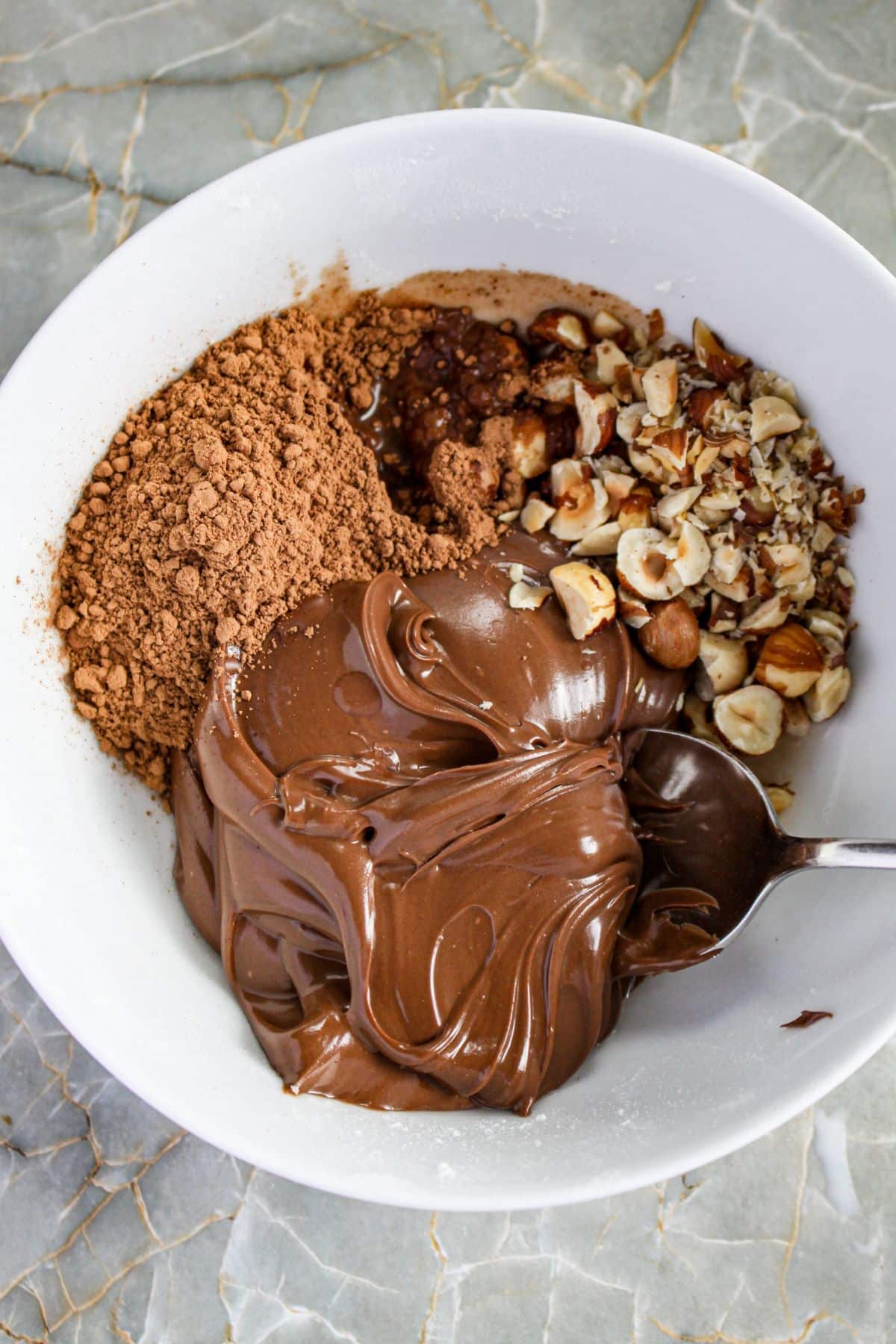 nutella, cocoa powder, and hazelnuts being mixed in a bowl.