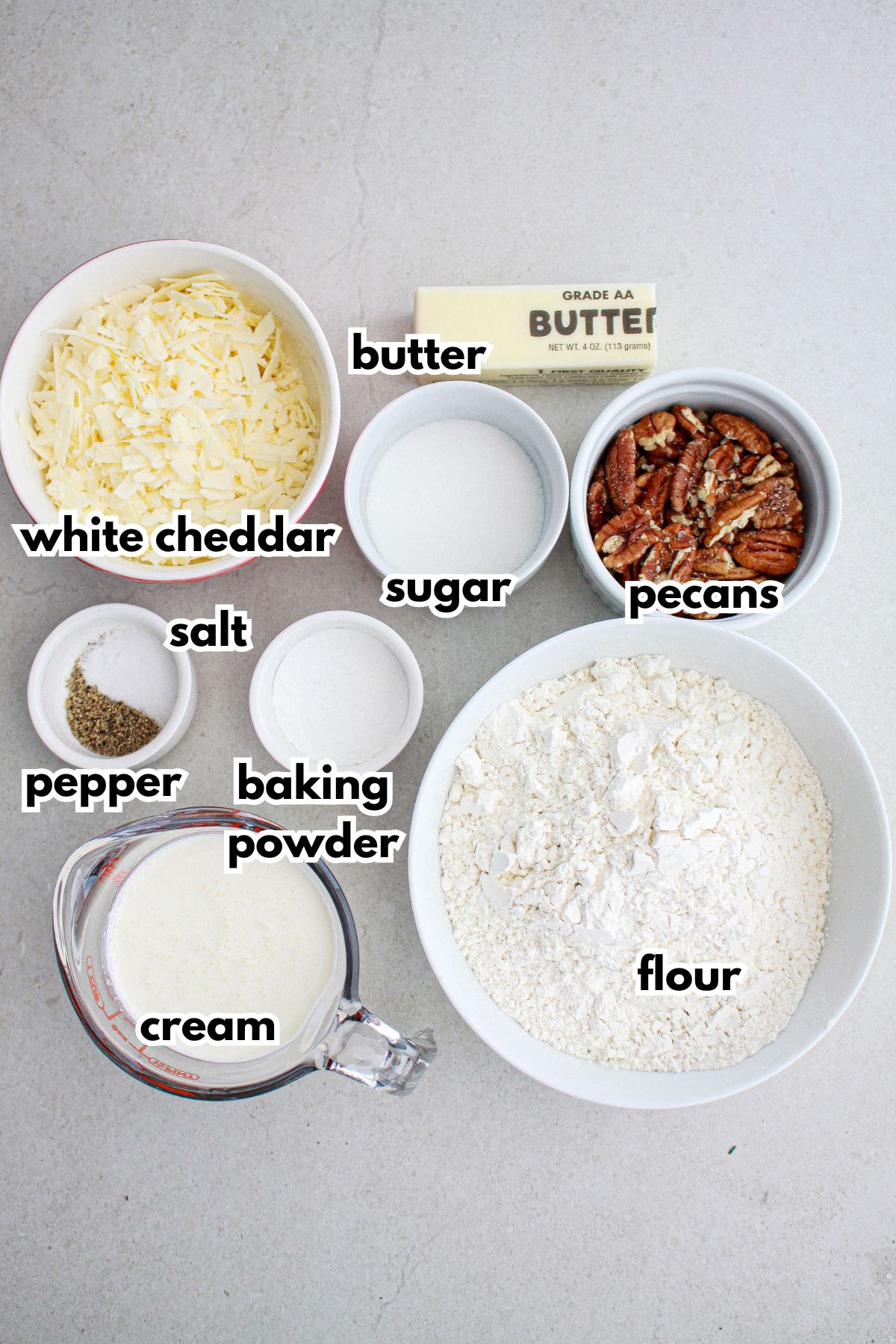 bowls of white cheddar cheese, butter, sugar, pecans, salt, pepper, baking powder, flour, and cream on a counter.