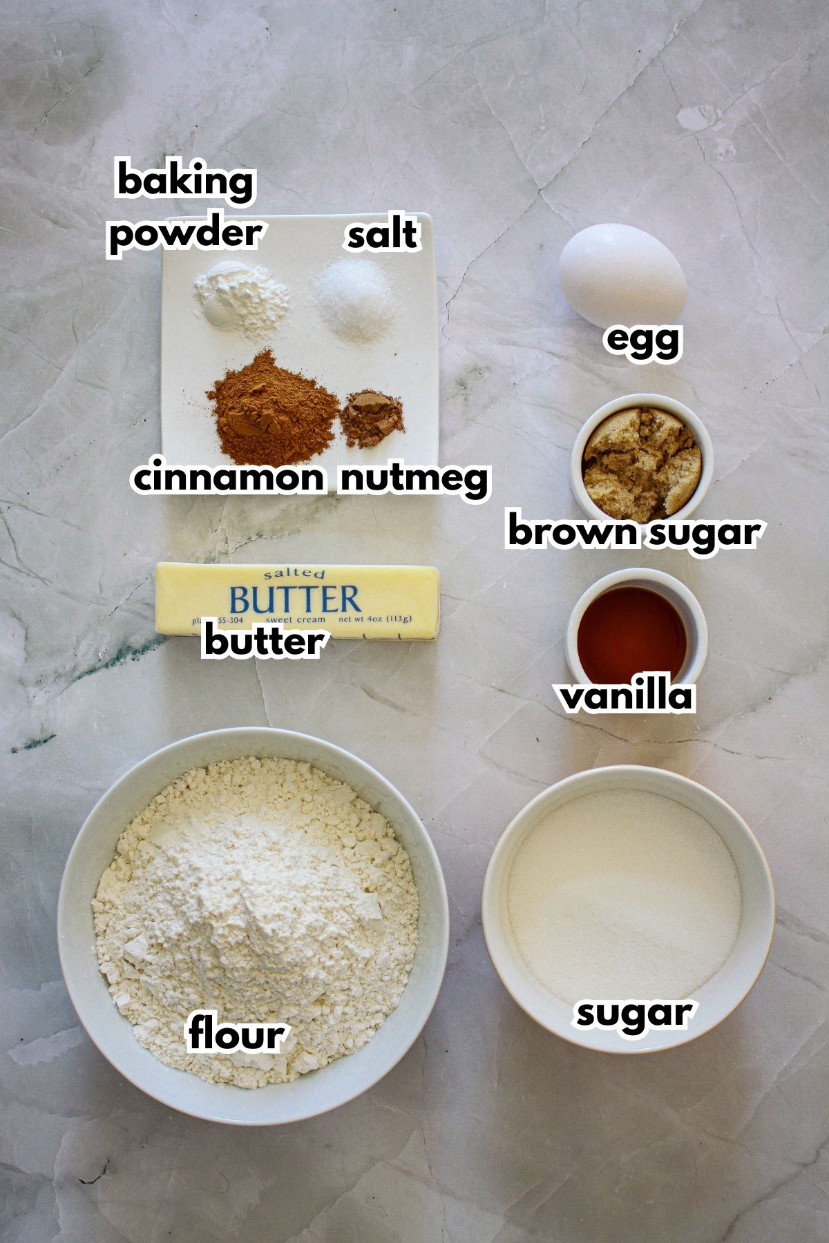 bowls of baking powder, salt, cinnamon, nutmeg, egg, brown sugar, butter, vanilla, flour, and sugar on a counter.