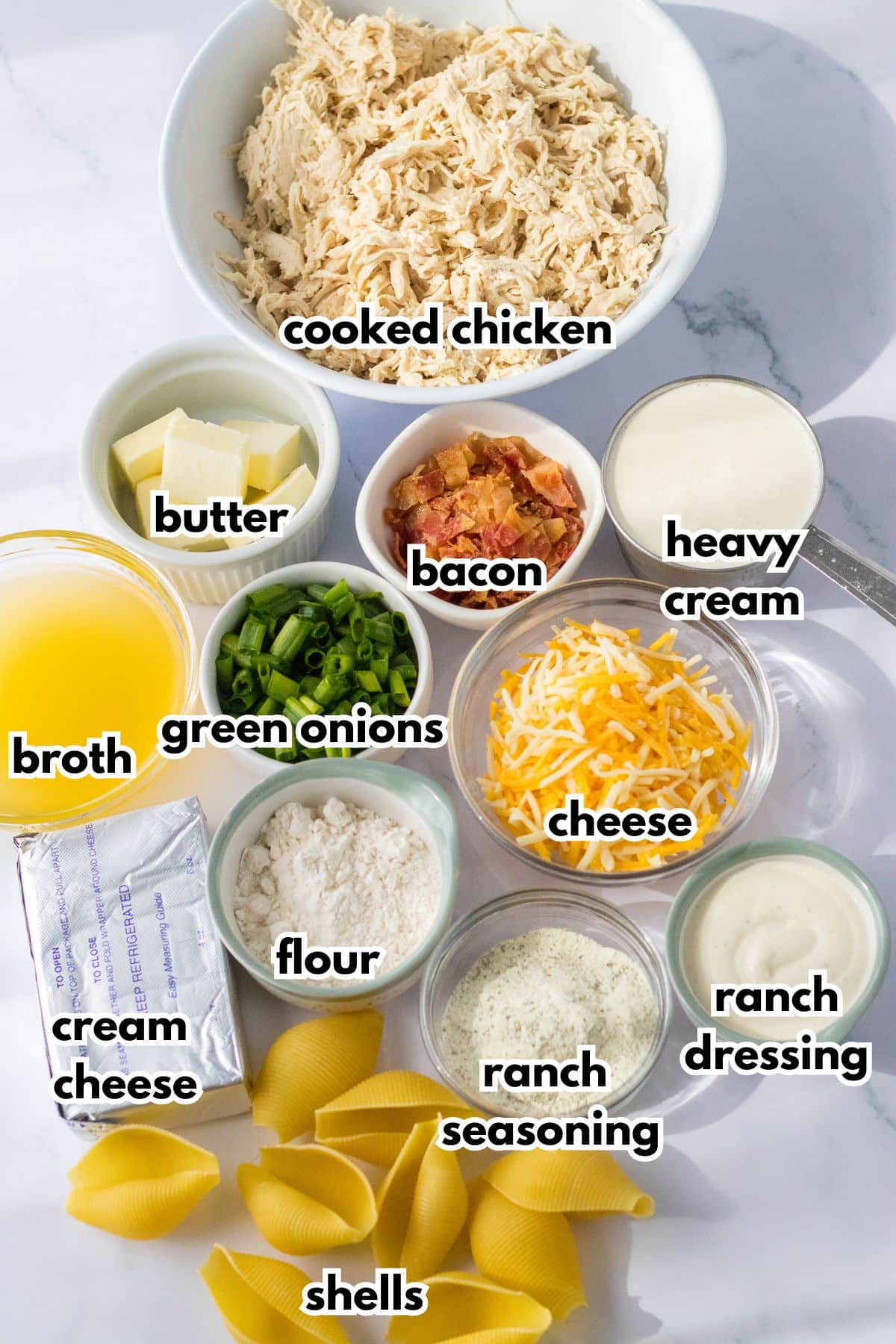 bowls of cooked chicken, butter, broth, bacon, green onions, heavy cream, cream cheese, flour, cheese, ranch seasoning, ranch dressing, and jumbo shells on a counter.