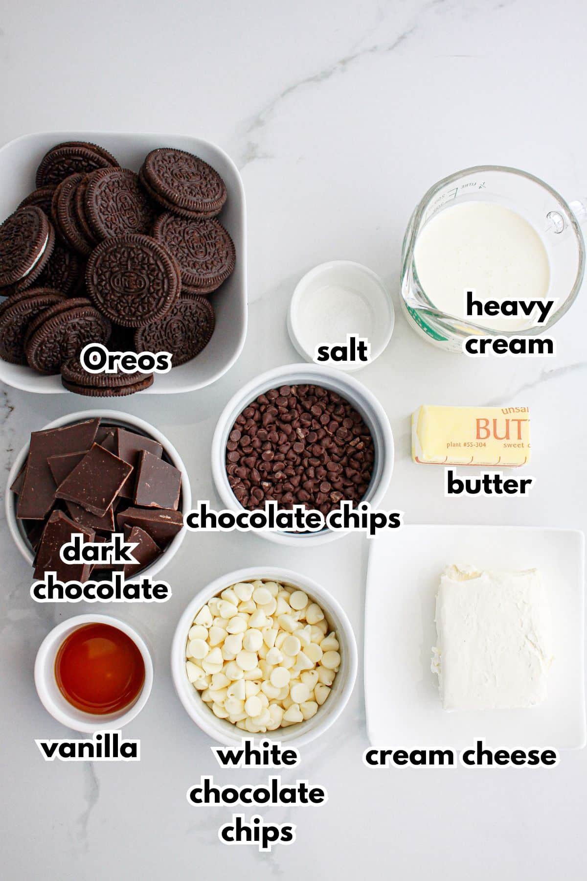 bowls of Oreos, salt, heavy cream, dark chocolate, chocolate chips, butter, vanilla, white chocolate, and cream cheese on a counter.