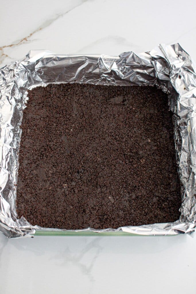 Oreo crust being pressed into a foil lined baking dish.