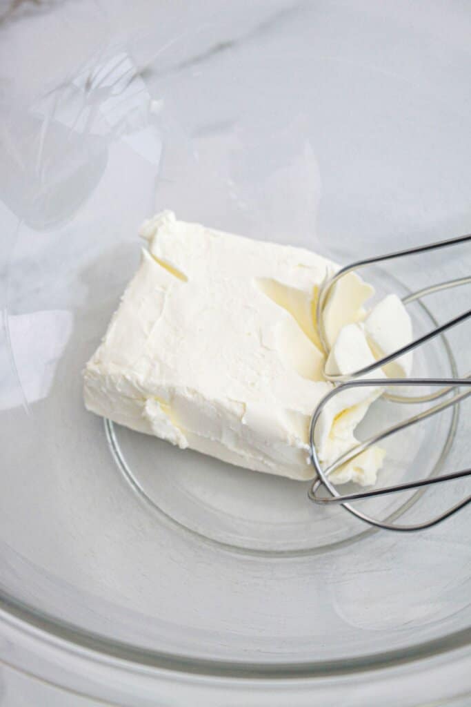 cream cheese being mixed with a hand mixer in a glass mixing bowl.