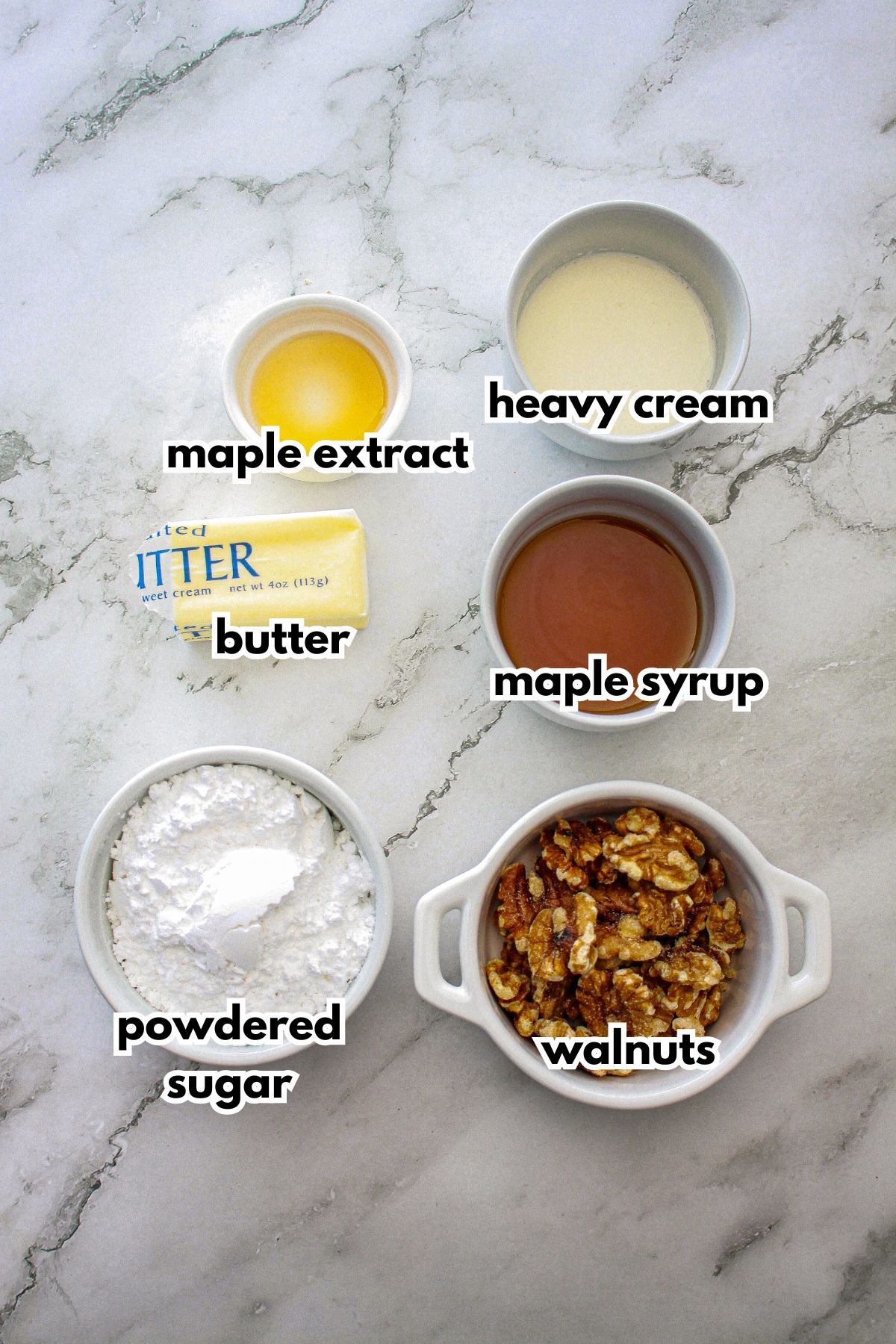 bowls of maple extract, butter, powdered sugar, heavy cream, maple syrup, and walnuts on a counter.