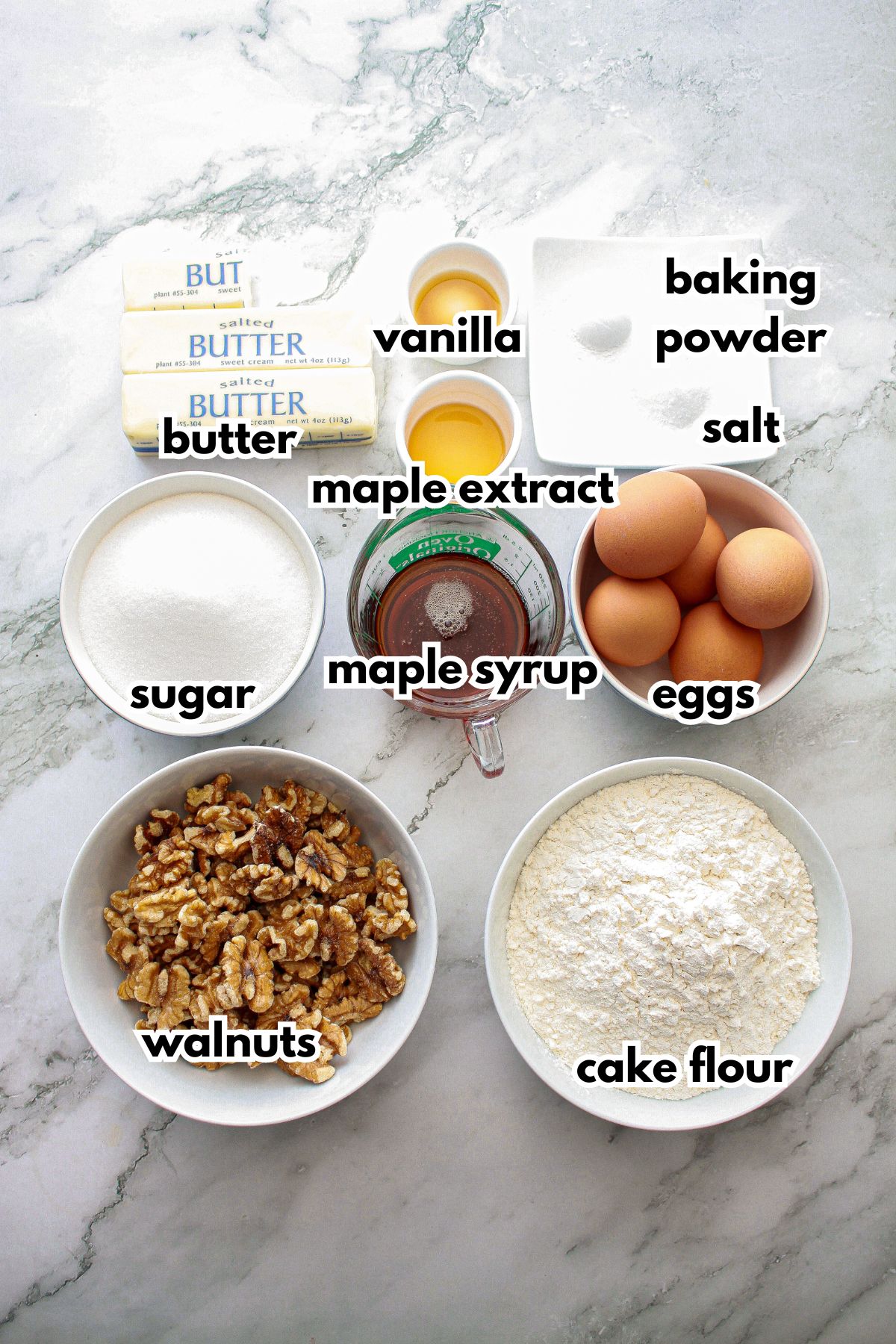 bowls of vanilla, maple extract, butter, baking powder, salt, sugar, maple syrup, eggs, walnuts, and cake flour on a counter.