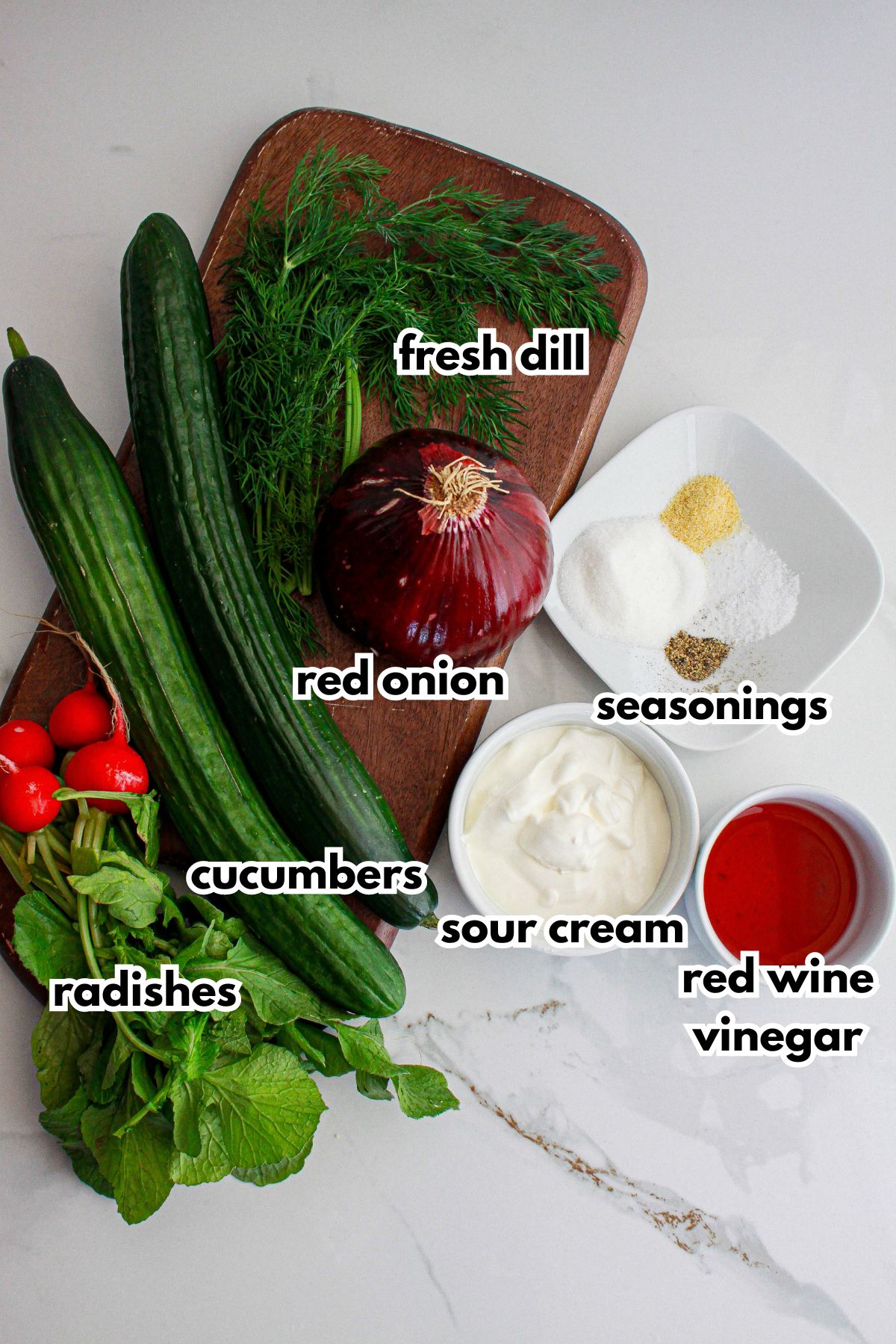 fresh dill, red onion, cucumbers, radishes, seasonings, sour cream, and red wine vinegar on a counter.