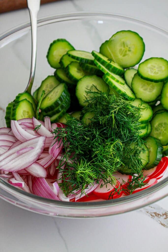 cucumbers, onions, radishes, dill, and dressing being combined in a glass mixing bowl.