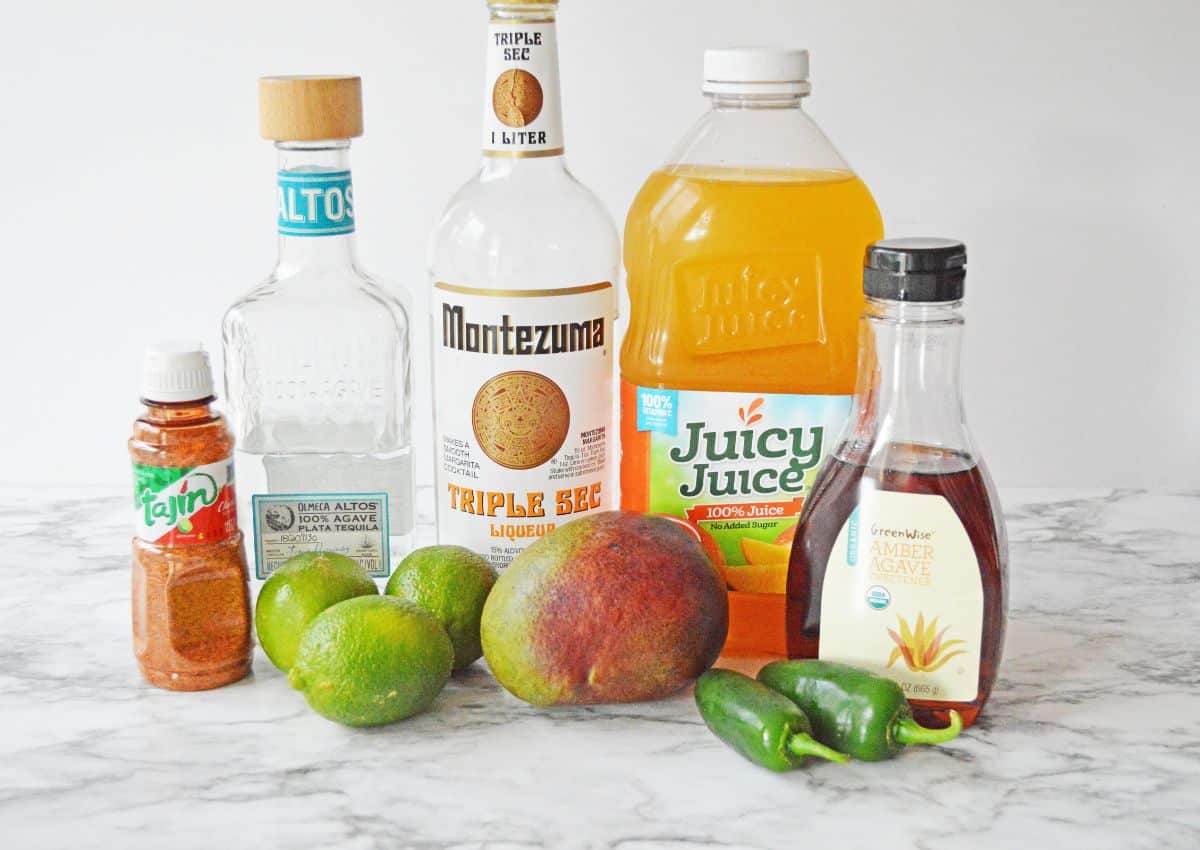 bowls of tajin, agave, triple sec, mango juice, limes, mangoes, jalapenos on a counter.