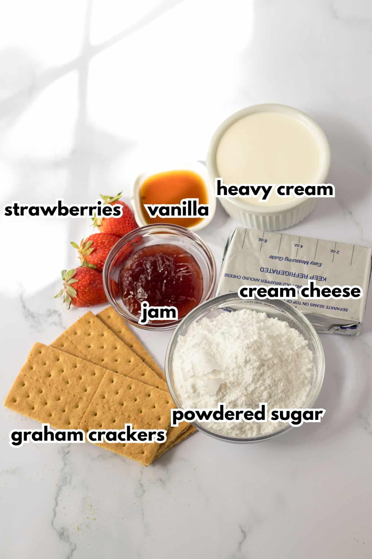 bowls of vanilla, heavy cream, jam. cream cheese, graham crackers, powdered sugar, and fresh strawberries on a counter.