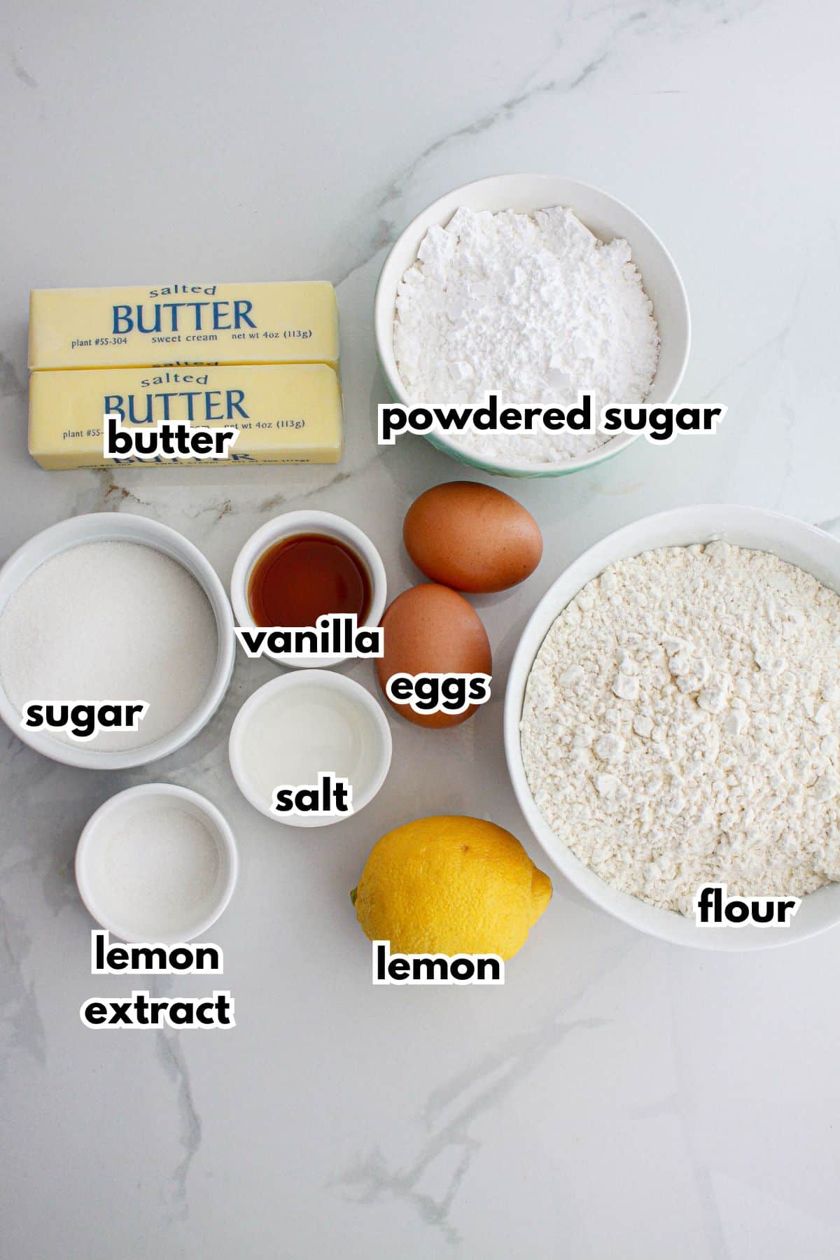 bowls of butter, powdered sugar, sugar, vanilla, eggs, lemon extract, salt, lemon, and flour on a counter.