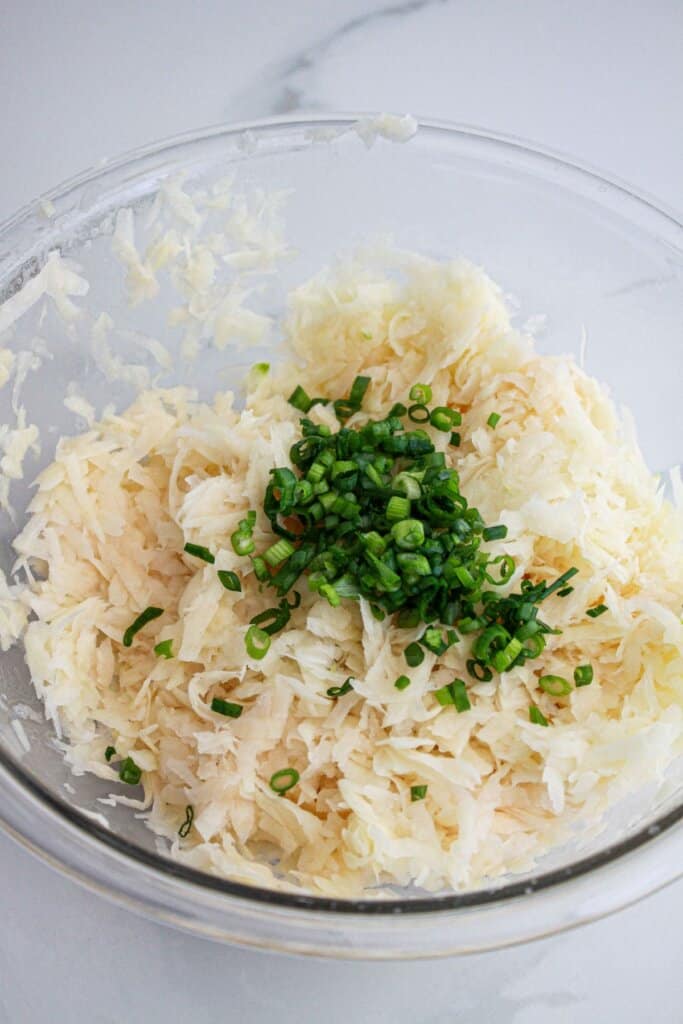 green onions being added to shredded potatoes and onions in a glass bowl.