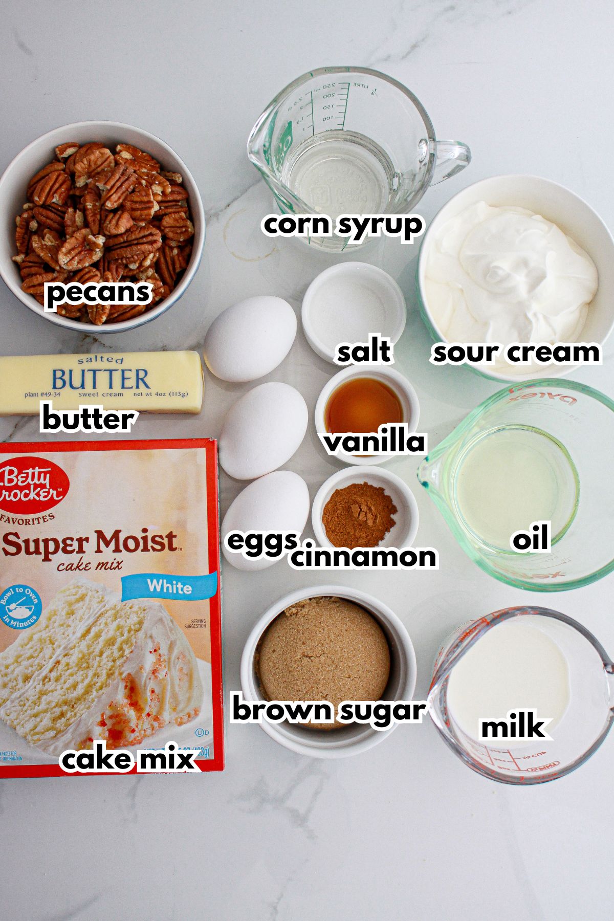 pecans, butter, cake mix, corn syrup, salt, vanilla, cinnamon, eggs, brown sugar, sour cream, oil and milk on a counter.
