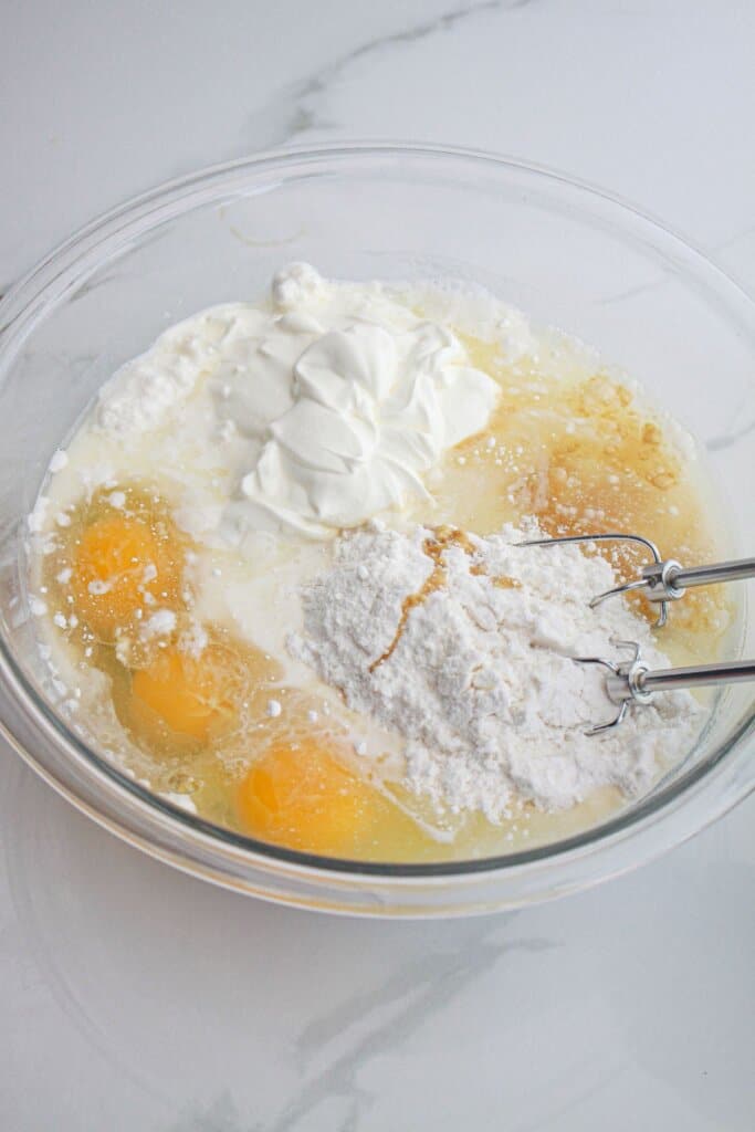cake mix, sour cream, and eggs being mixed together with a hand mixer in a glass mixing bowl.