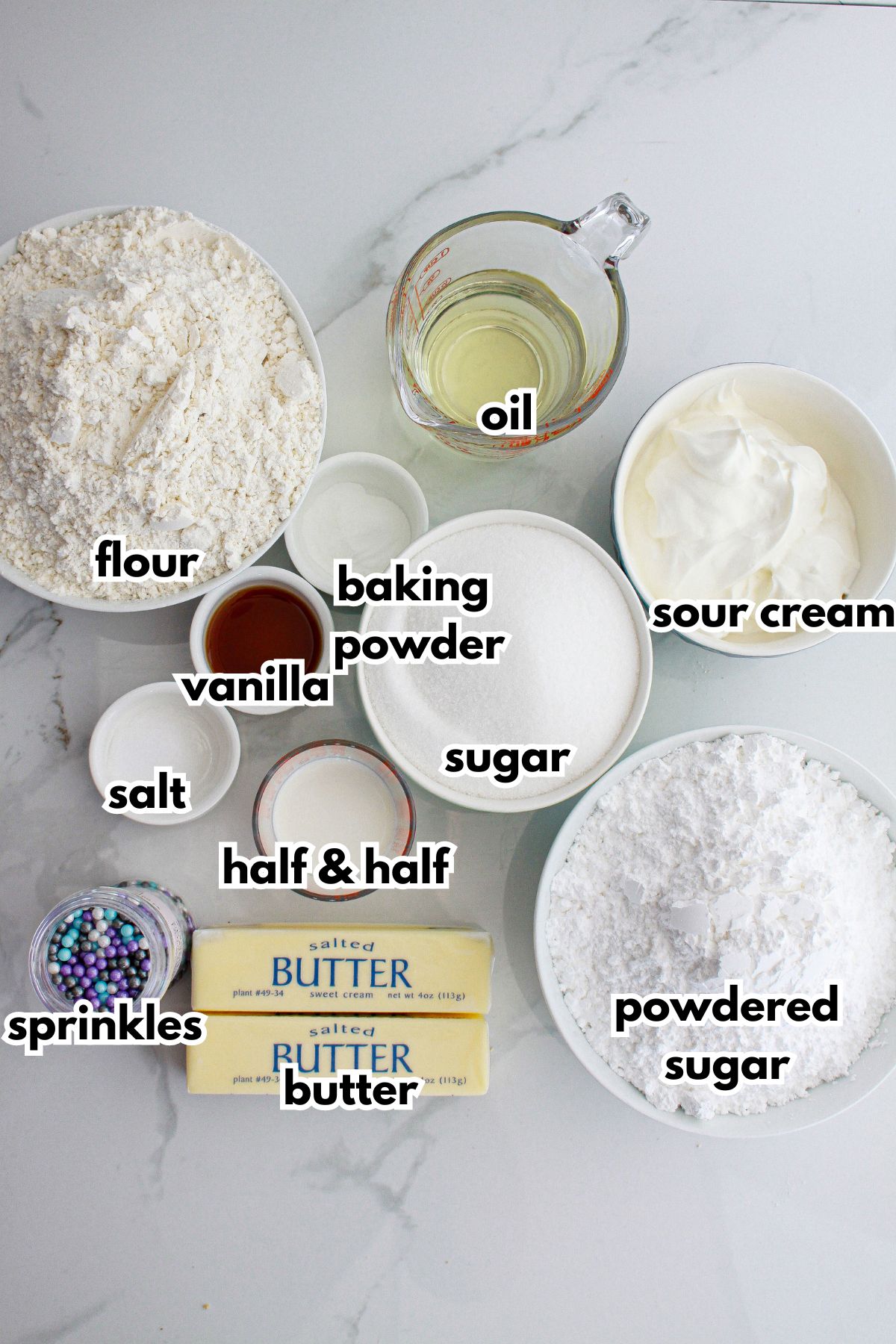 bowls of flour, baking powder, oil, vanilla, salt, sugar, sour cream, half and half, sprinkles, butter, and powdered sugar on a counter.