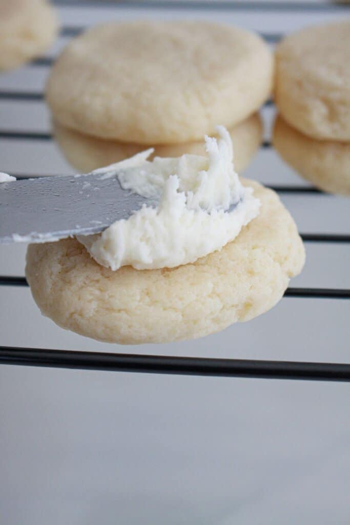 frosting being spread on top of a sugar cookie on a cooling rack.