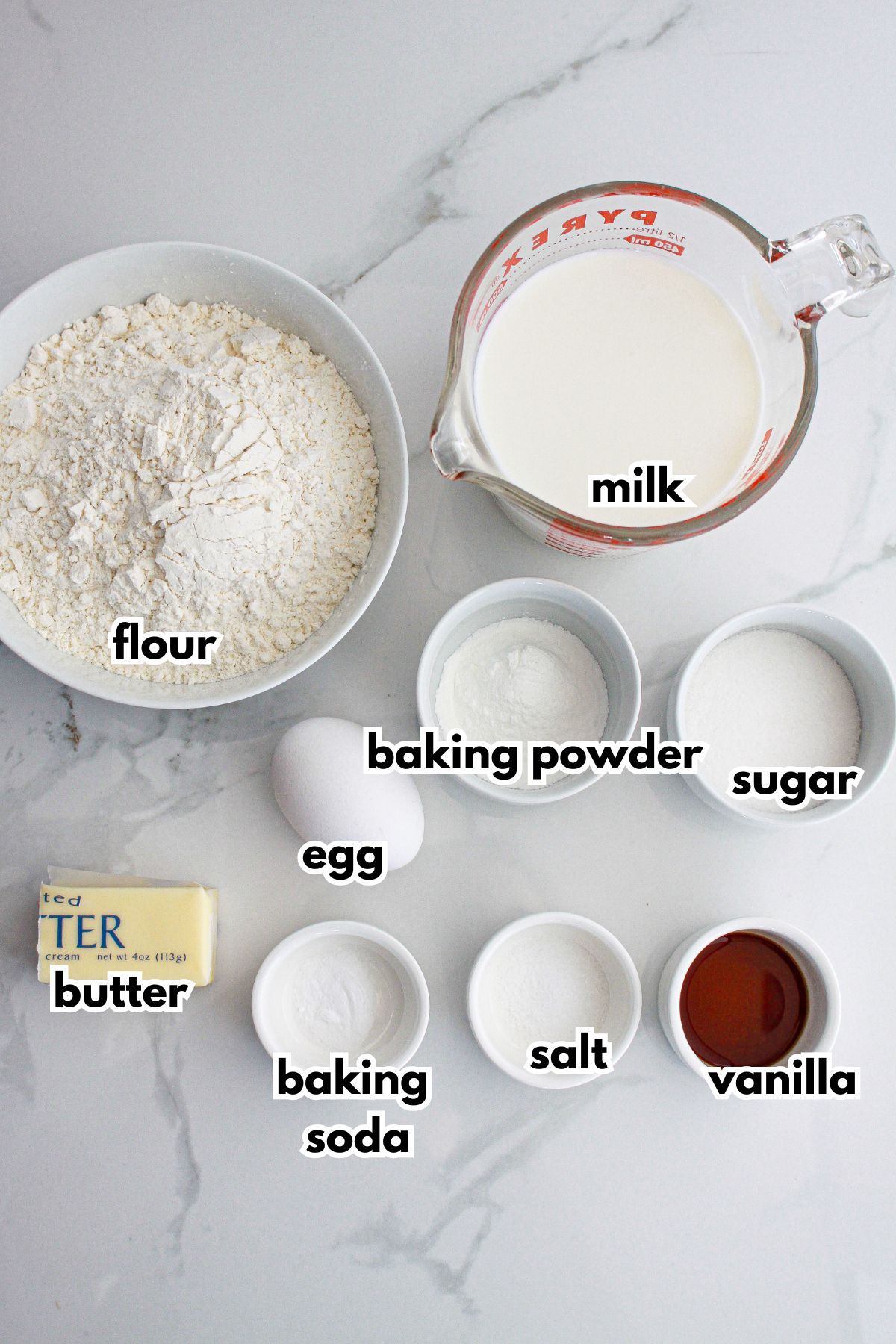 bowls of flour, milk, baking powder, sugar, egg, butter, baking soda, salt, and vanilla on a counter.