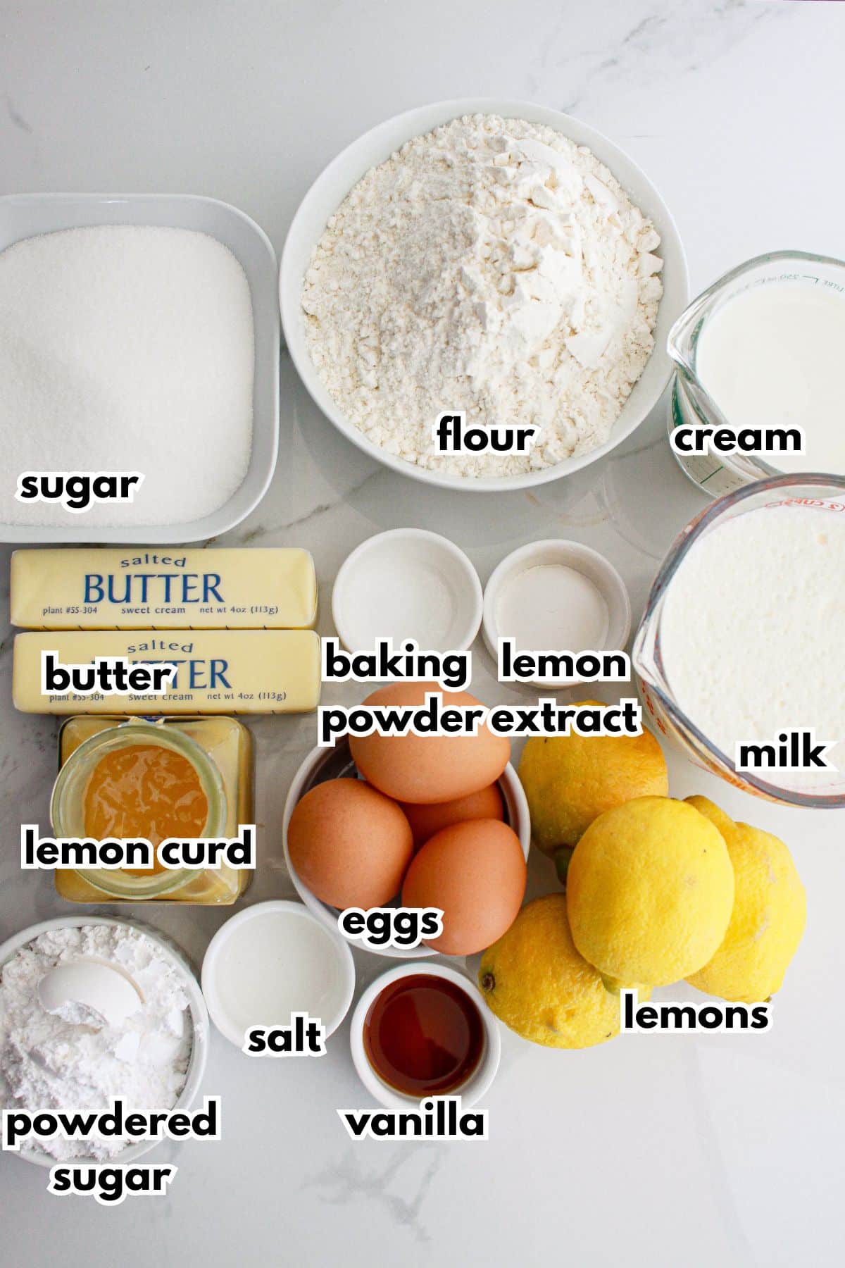 bowls of sugar, flour, cream, butter, baking powder, lemon extract, milk, lemon curd, eggs, lemons, powdered sugar, salt, and vanilla on a counter.