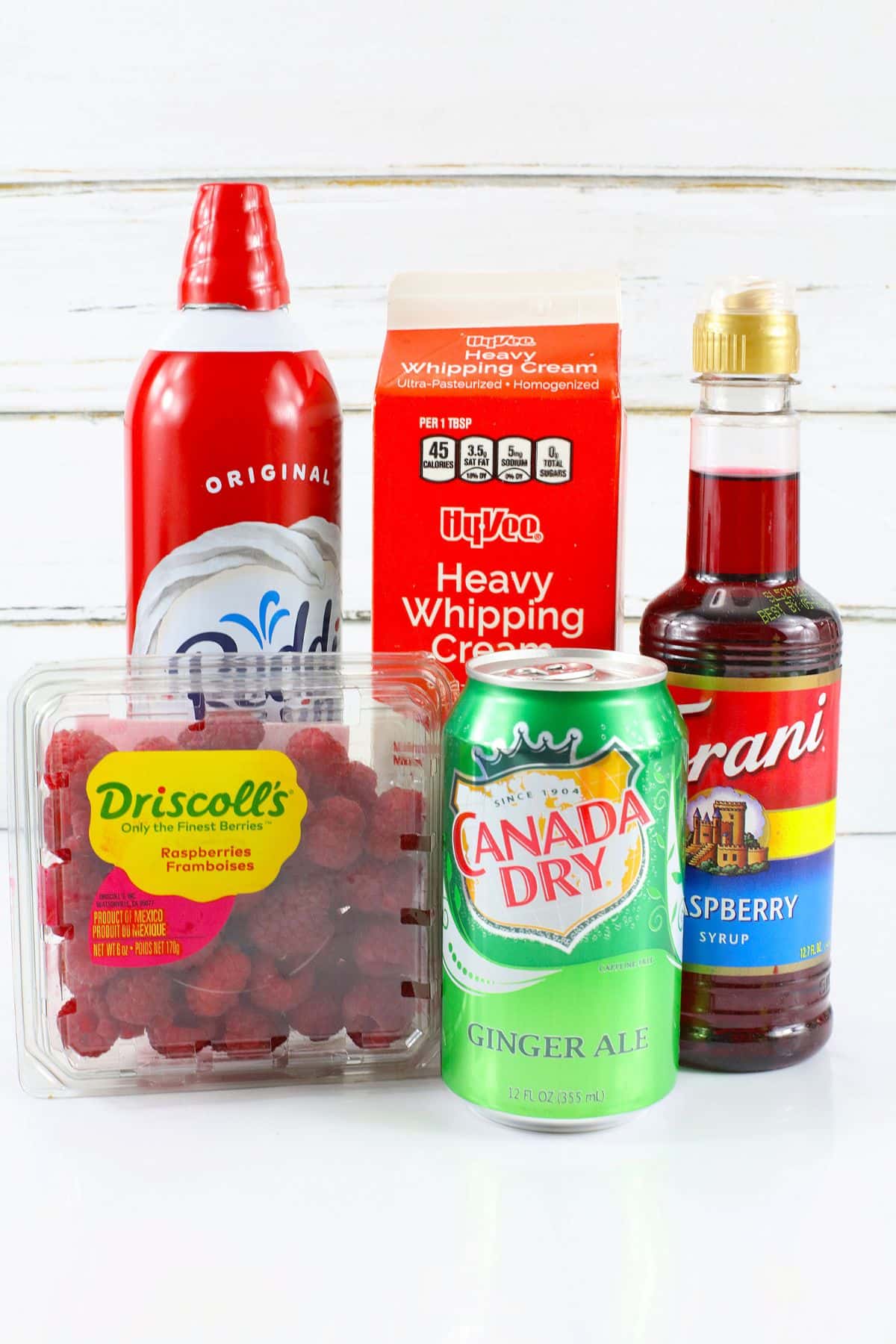 raspberries, heavy cream, whipped cream. ginger ale, and raspberry syrup on a counter.