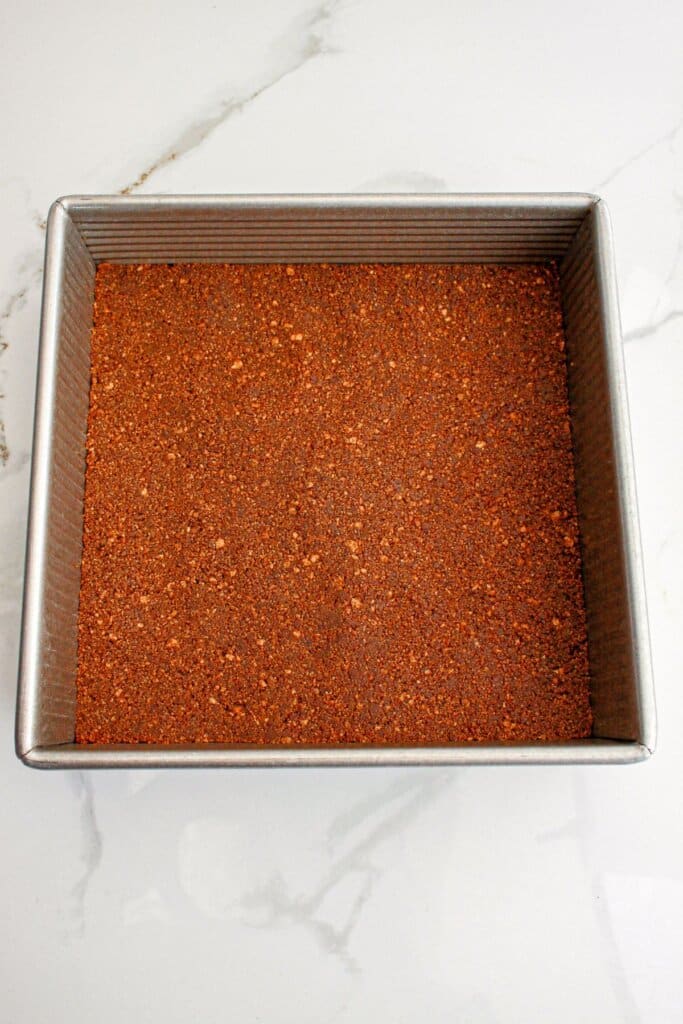 chocolate graham cracker crust being pressed into the bottom of a baking pan.