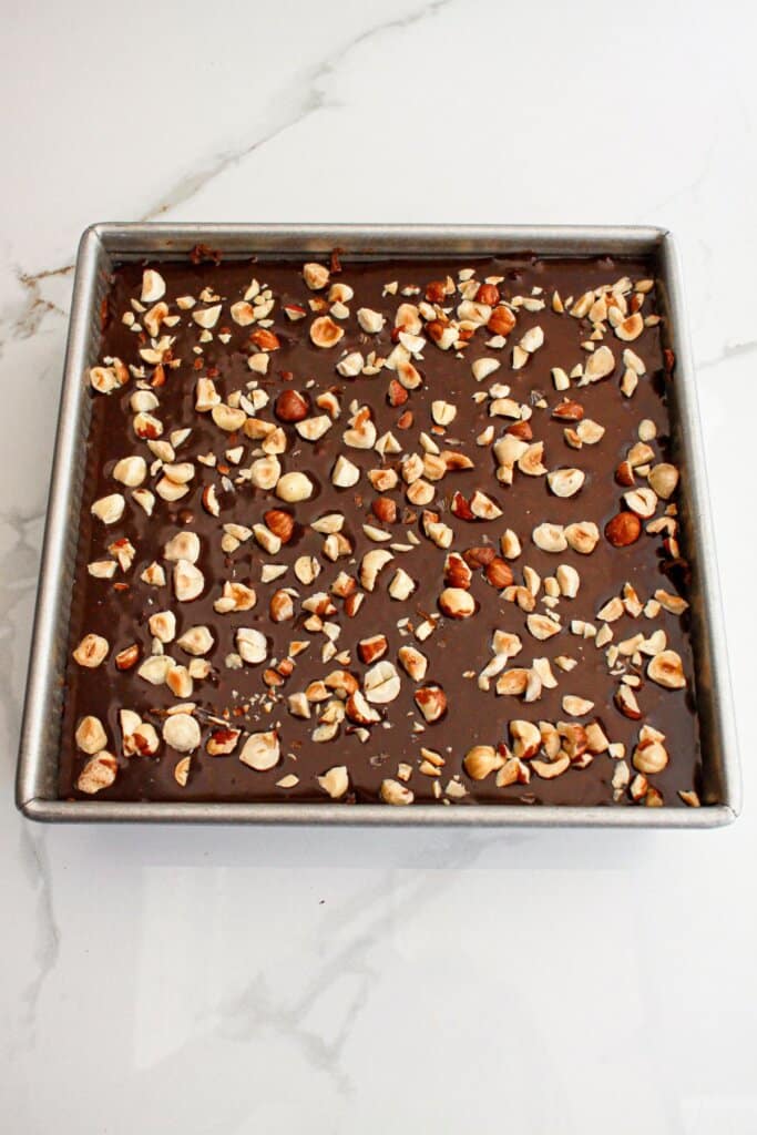 hazelnuts being sprinkled on top of cheesecake bars in a baking dish.