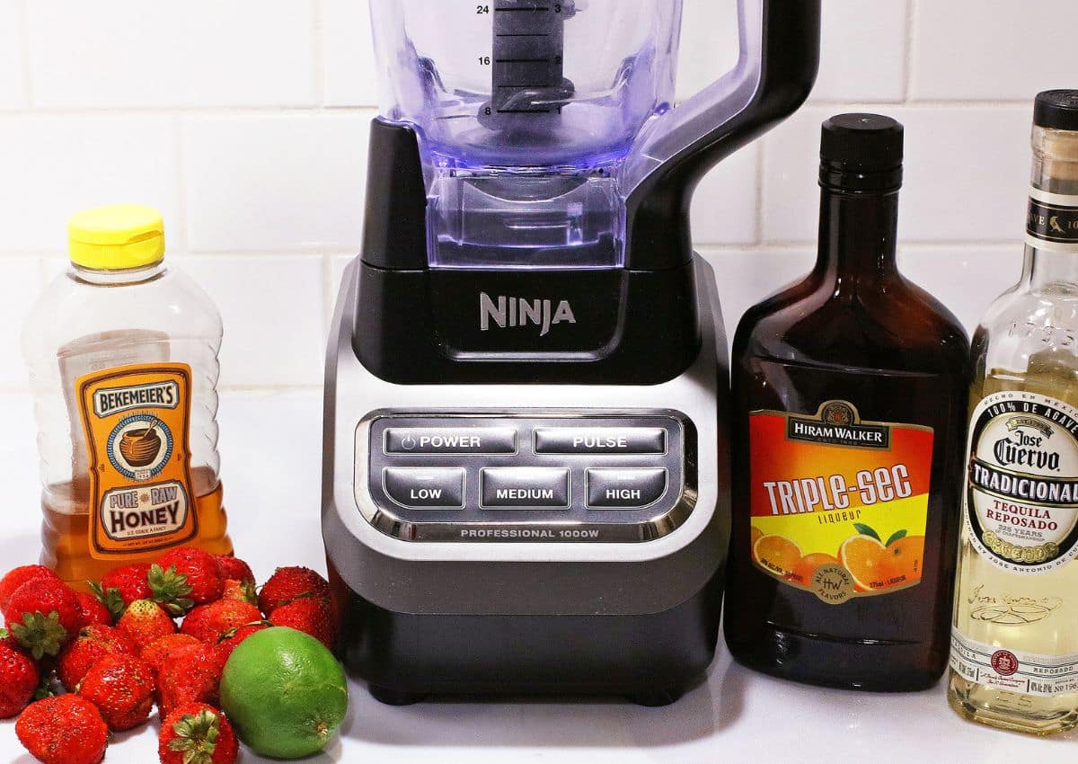 bottles of honey, triple sec, tequila, limes, and strawberries and a blender on a counter.