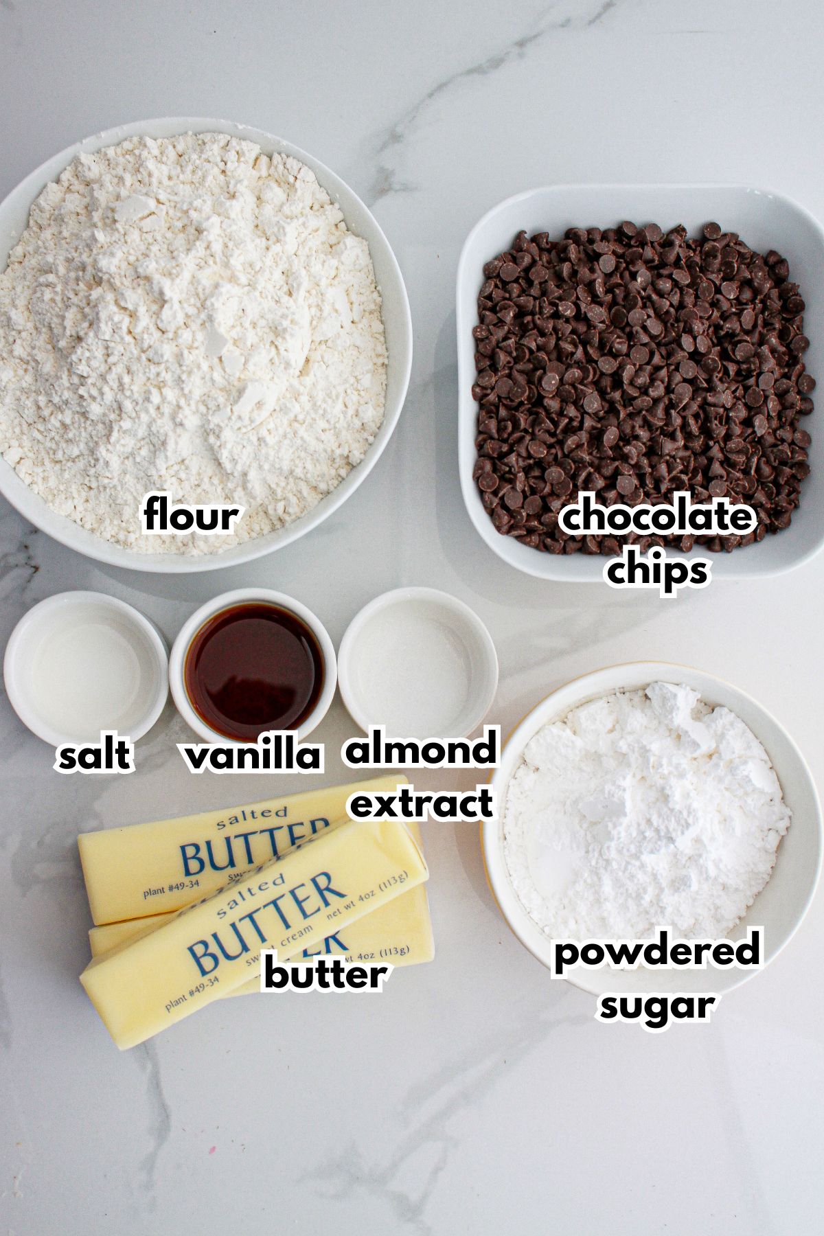 bowls of flour, chocolate chips, salt, vanilla, almond extract, butter, and powdered sugar on a counter.