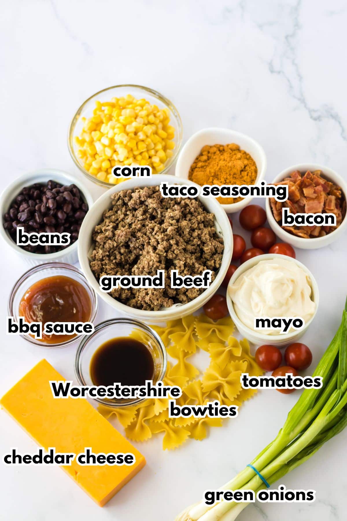 bowls of corn, taco seasoning, bacon, beans, bbq sauce, ground beef, worcestershire, cheddar cheese, bowties, mayo, and green onions on a counter.