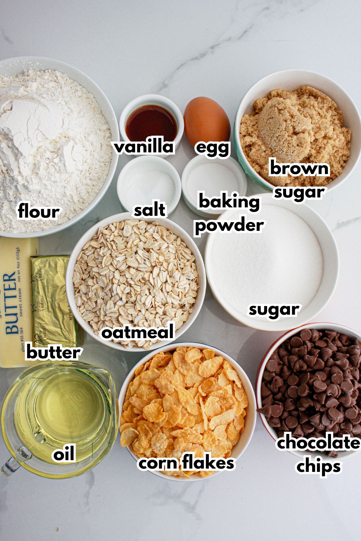 bowls of flour, salt, baking powder, vanilla, egg, brown sugar, butter, oatmeal, sugar, oil, corn flakes, and chocolate chips on a counter.