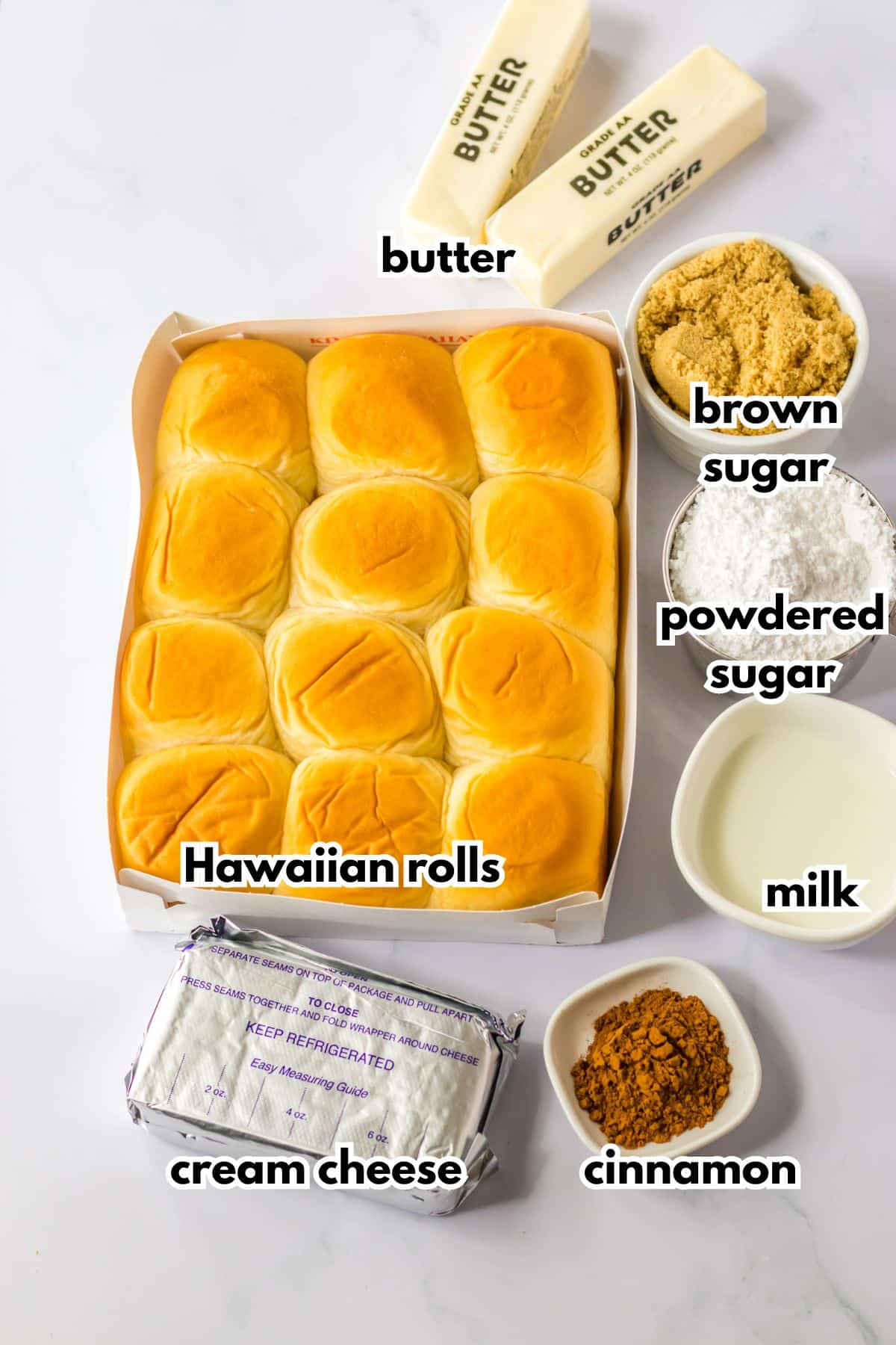 box of hawaiian rolls, sticks of butter, block of carem cheese, powdered sugar, brown sugar, milk and cinnamon on a counter.