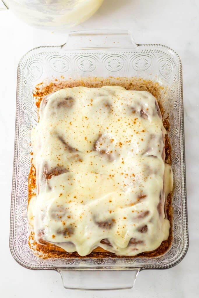 cream cheese frosting being spread on top of rolls in a baking dish.