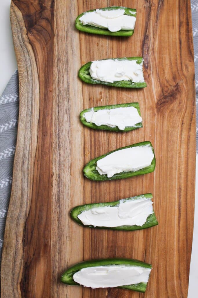 cream cheese being spread inside of peppers on a wooden cutting board.
