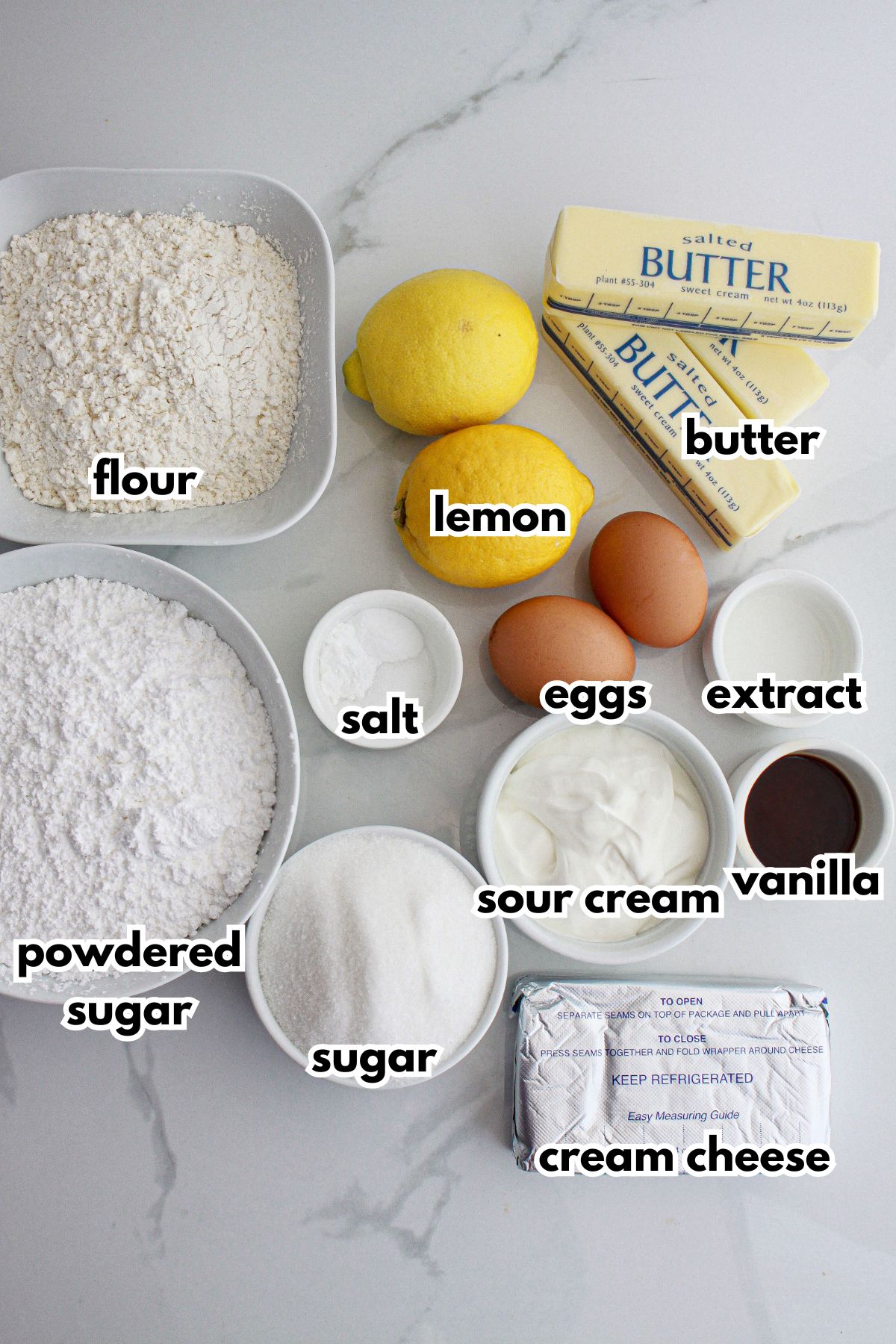 bowls of flour, lemon, butter, salt, powdered sugar, eggs, lemon extract, vanilla, sour crea, sugar, and cream cheese on a counter.