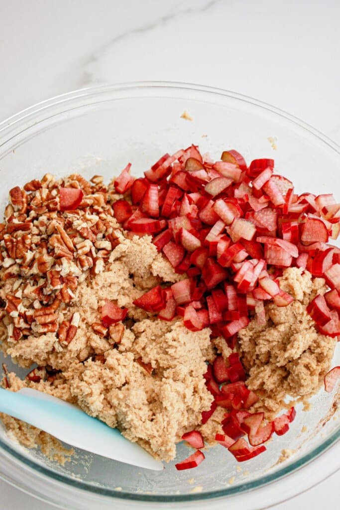 diced rhubarb and pecans being added to cookie dough in a glass bowl.